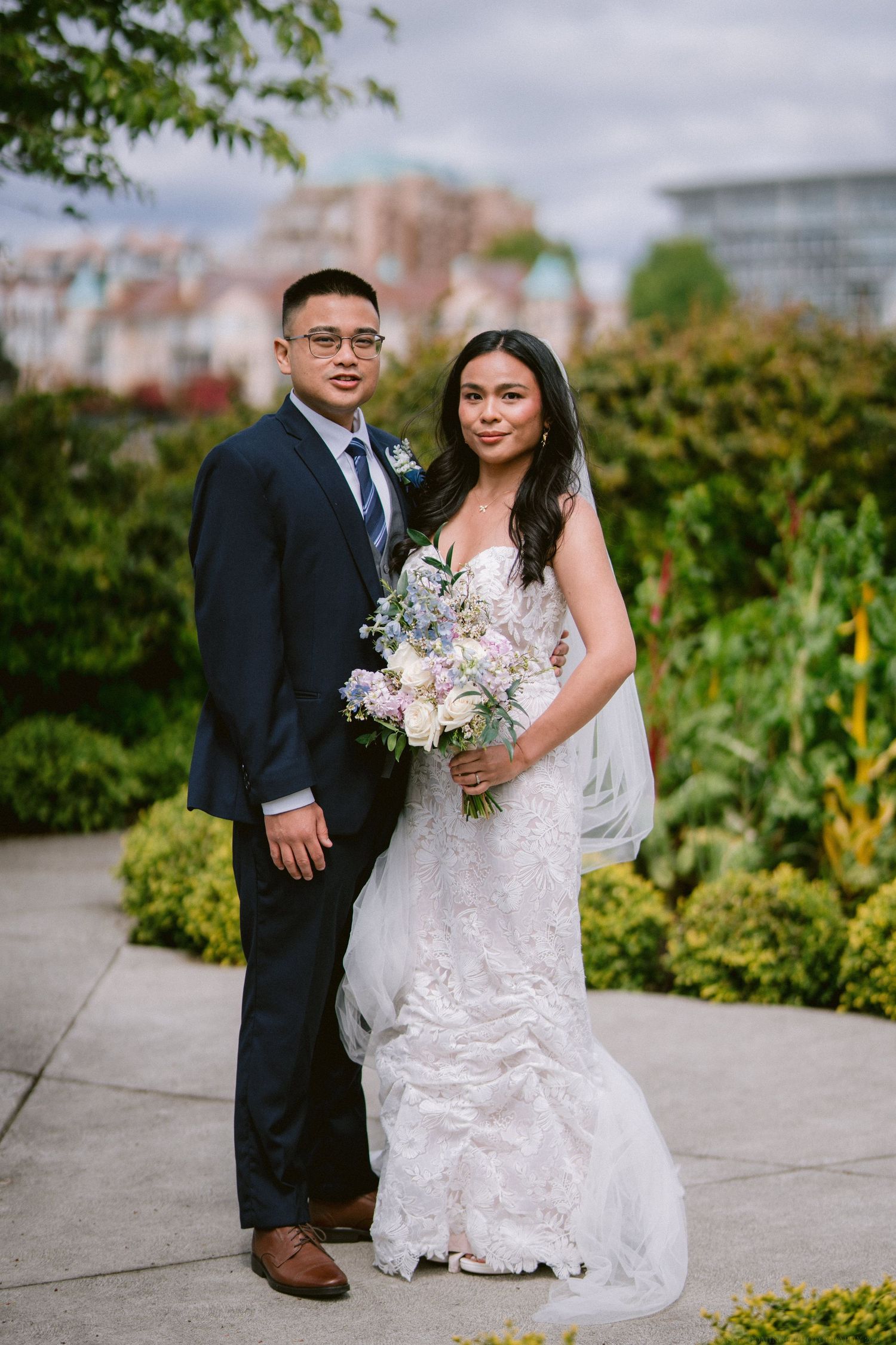 Wedding couple poses in a garden setting with lush greenery and urban backdrop.