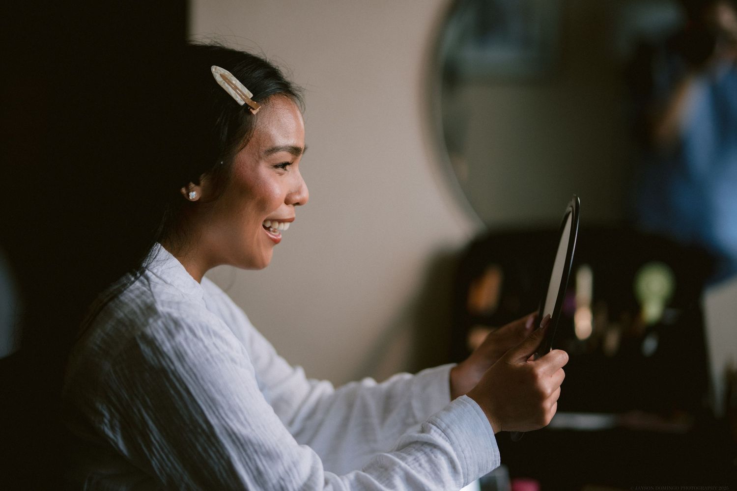 A person in a white robe smiles while looking at a mobile device during wedding preparations.