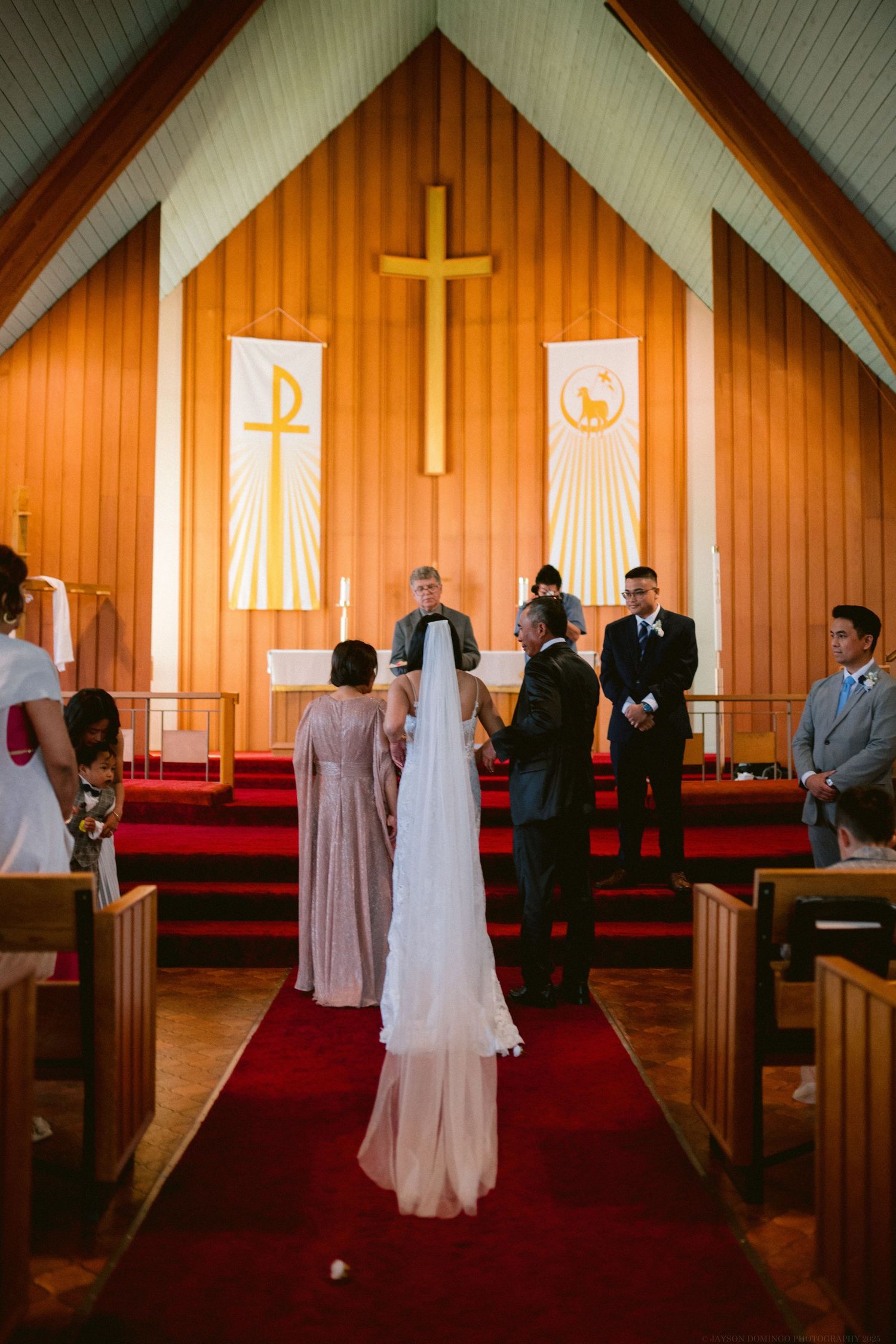 A traditional wedding ceremony takes place in a church sanctuary with wooden walls and a golden cross at the altar.