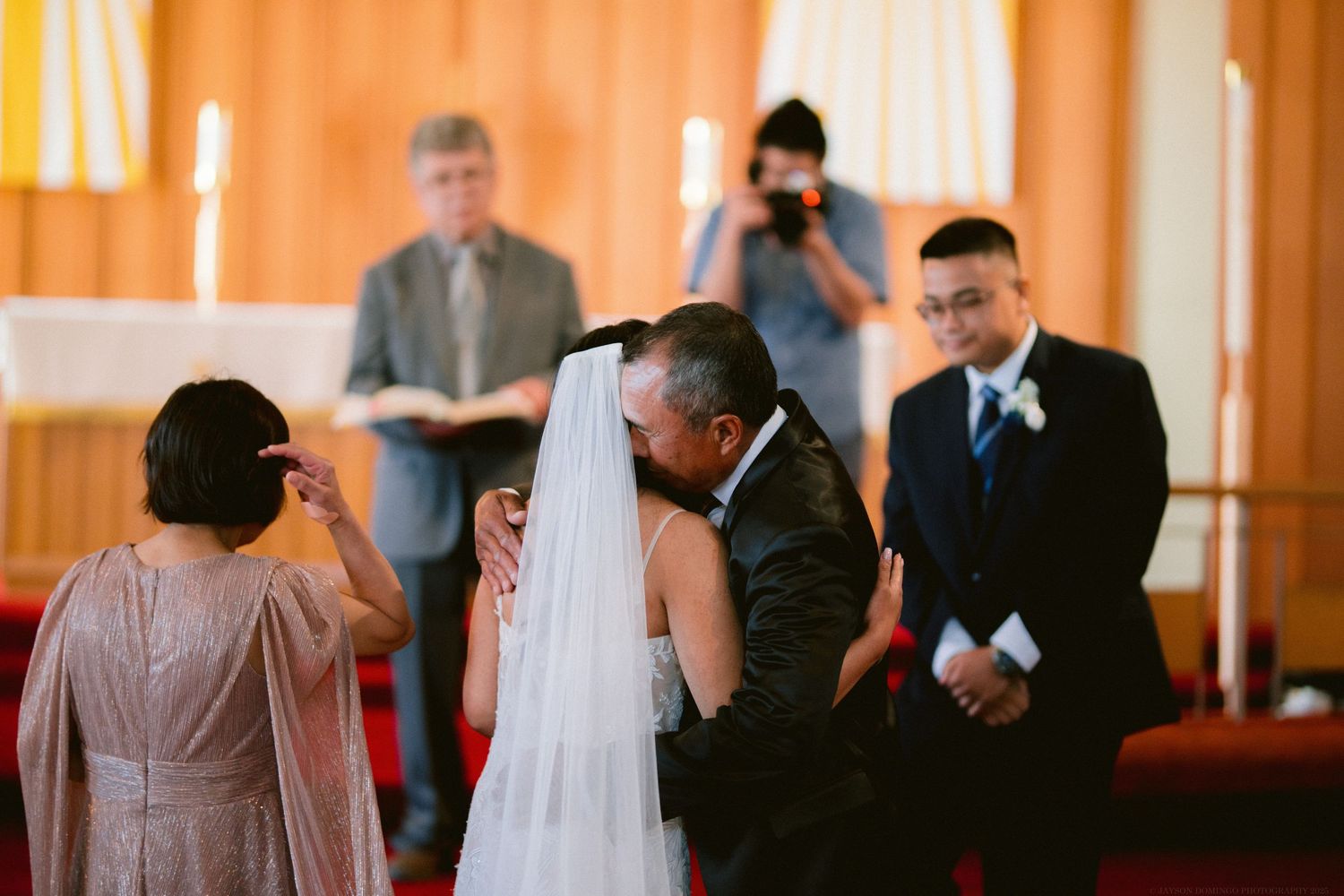 An emotional wedding ceremony takes place in a church with orange drapery and stained glass windows.