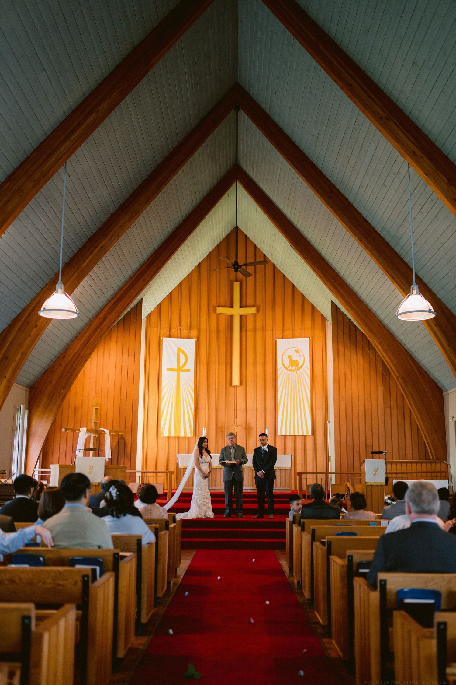 Light flows through a vaulted wooden church sanctuary with red carpet during a wedding ceremony.