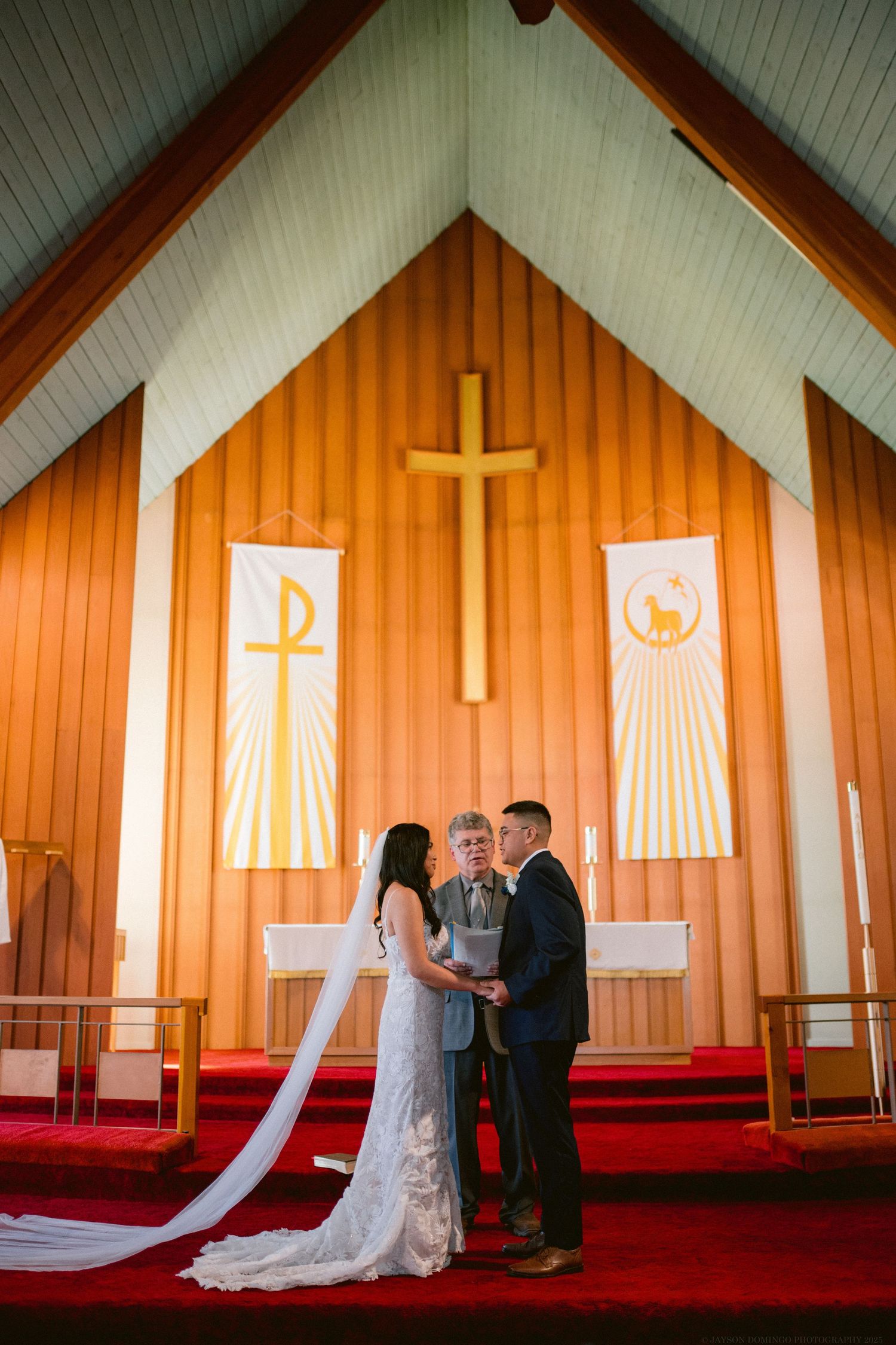 A wedding ceremony takes place in a church with orange wood walls and religious banners.