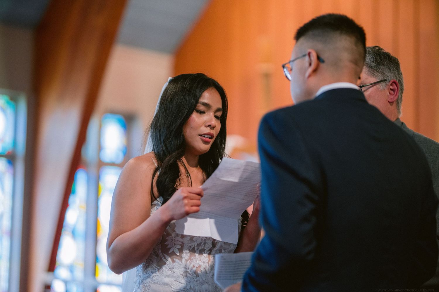 A bride in a white gown reads vows during an indoor wedding ceremony with stained glass windows.