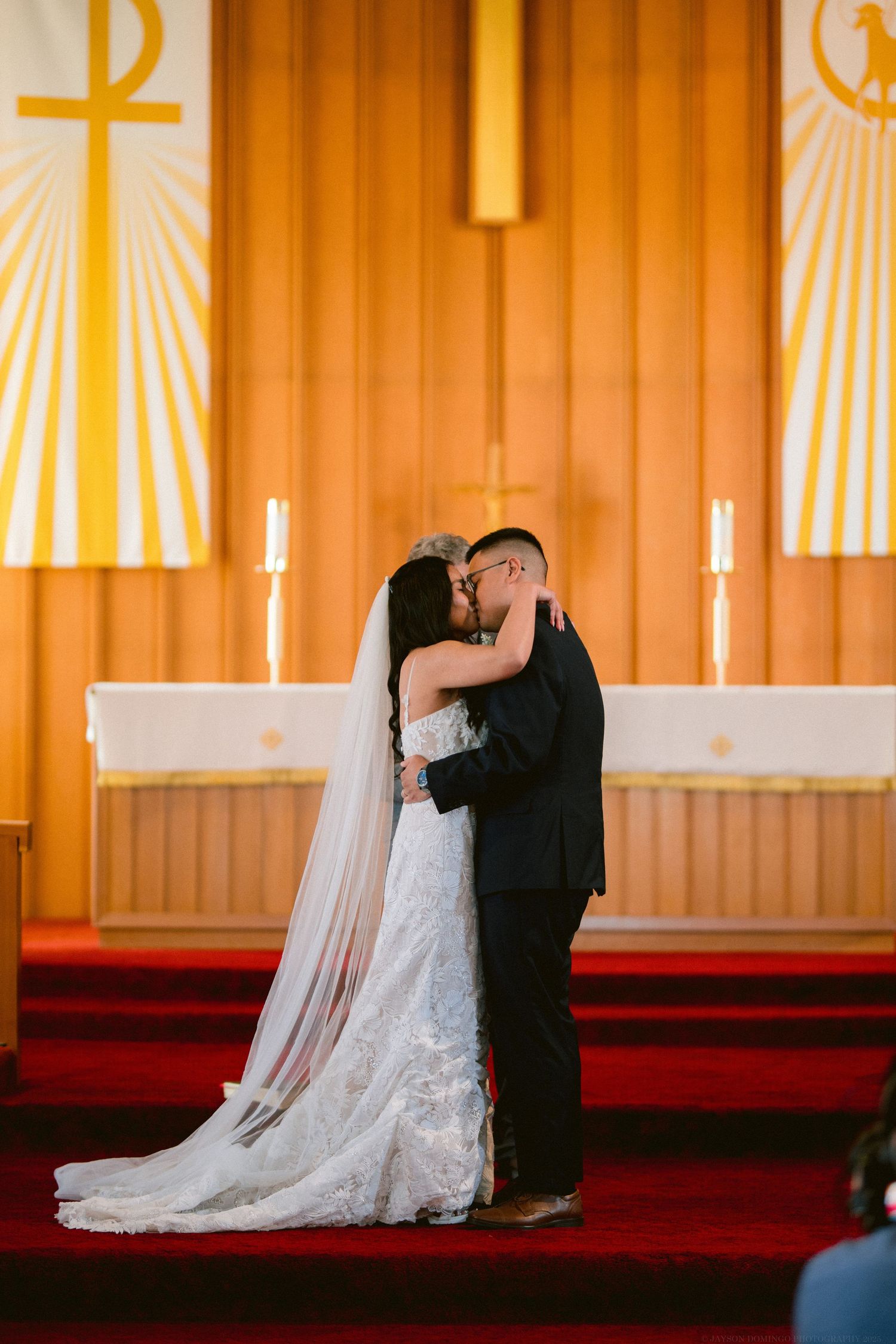 Newlyweds share their first kiss as a married couple at the altar with golden religious banners behind them.