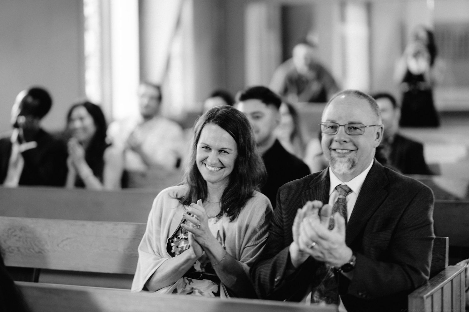 Wedding guests smile and applaud during the ceremony in church pews.