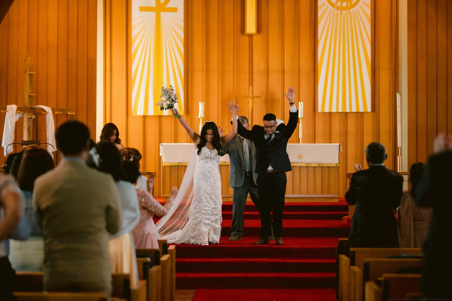 Newlyweds celebrate with raised arms at the altar of a church decorated with golden banners.