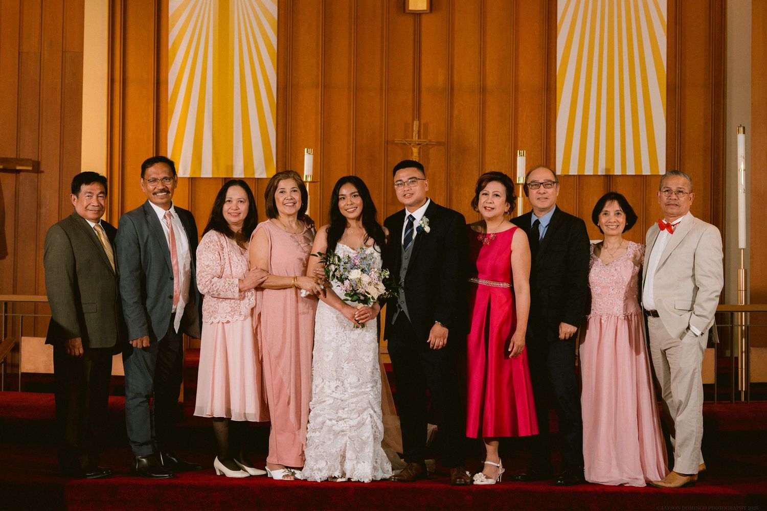 Wedding party lineup in pink and formal attire posing in front of wooden church walls.