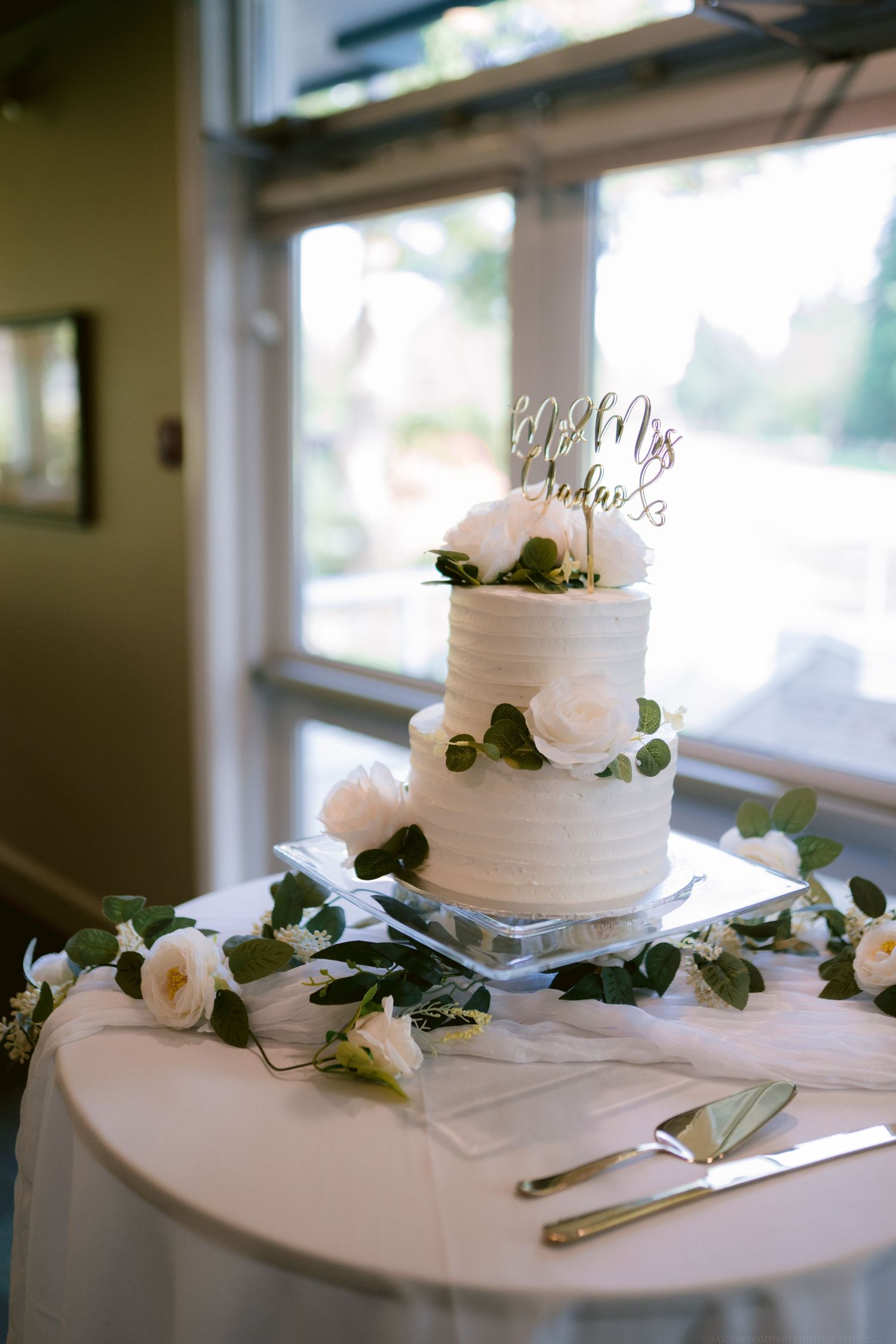 An elegant white wedding cake decorated with roses and greenery sits on a round table.