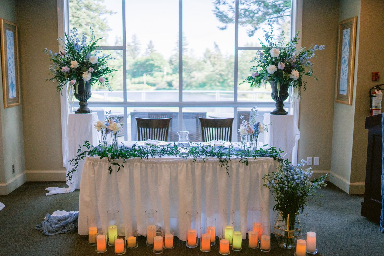 Wedding head table decorated with white linens, candles, and tall floral arrangements near windows.
