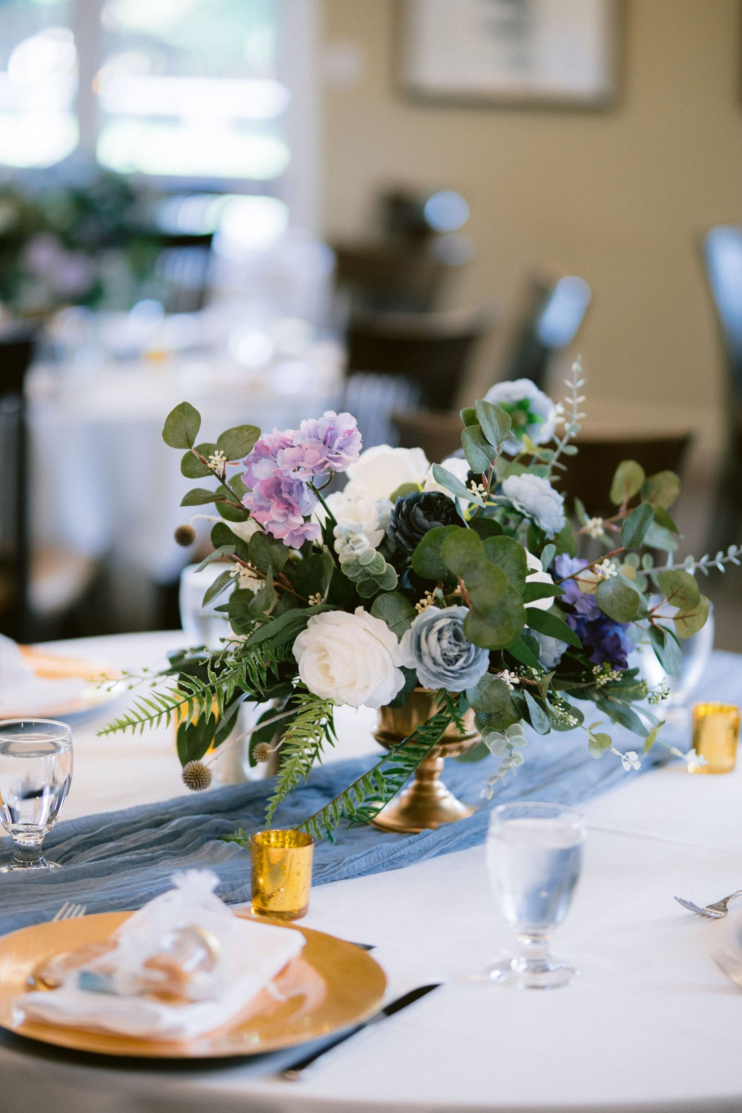 A romantic centerpiece featuring purple and white flowers with eucalyptus in a gold pedestal vase on a white tablecloth.