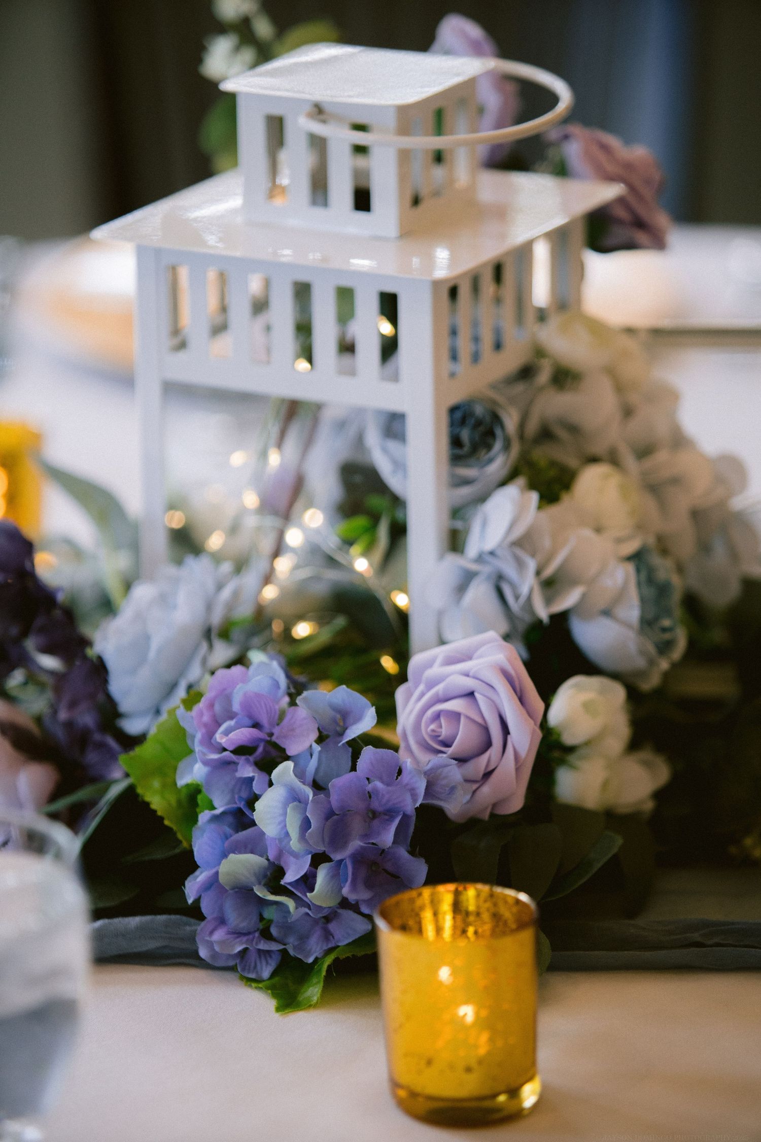 White lantern centerpiece surrounded by purple hydrangeas, roses, and amber votives on reception table.