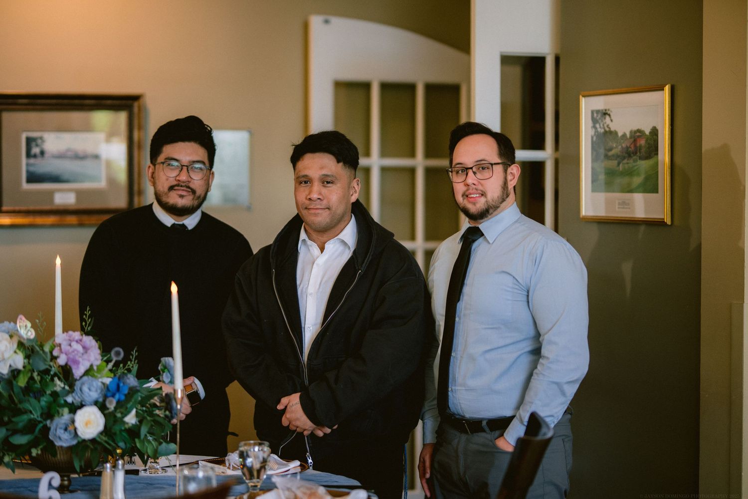 Three men in business attire stand together beside a floral arrangement in an office hallway with framed artwork.