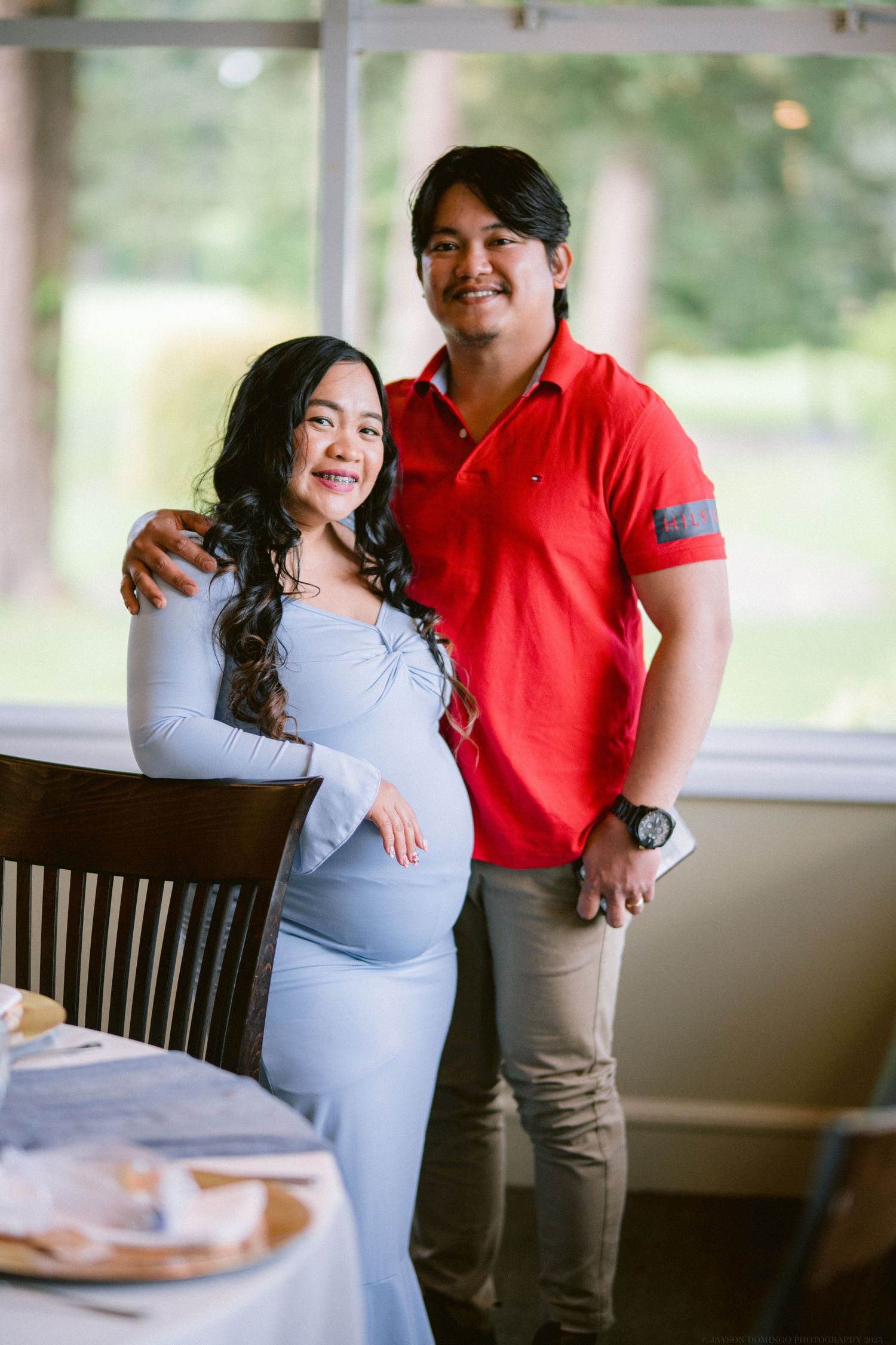 Couple poses together by window, with person wearing a light blue maternity dress.
