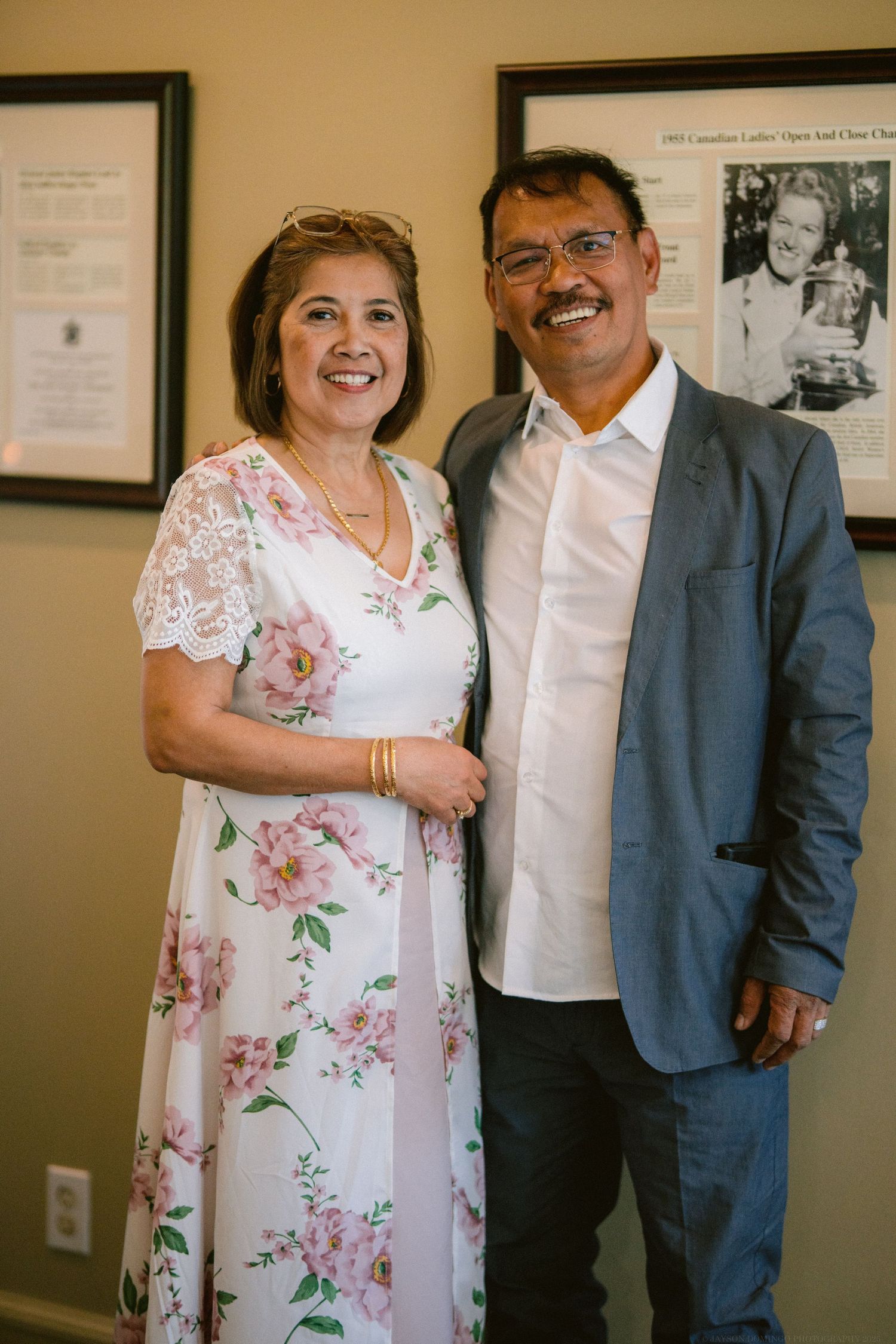 A couple in formal attire pose together, with the woman wearing a floral white dress and the man in a gray suit.