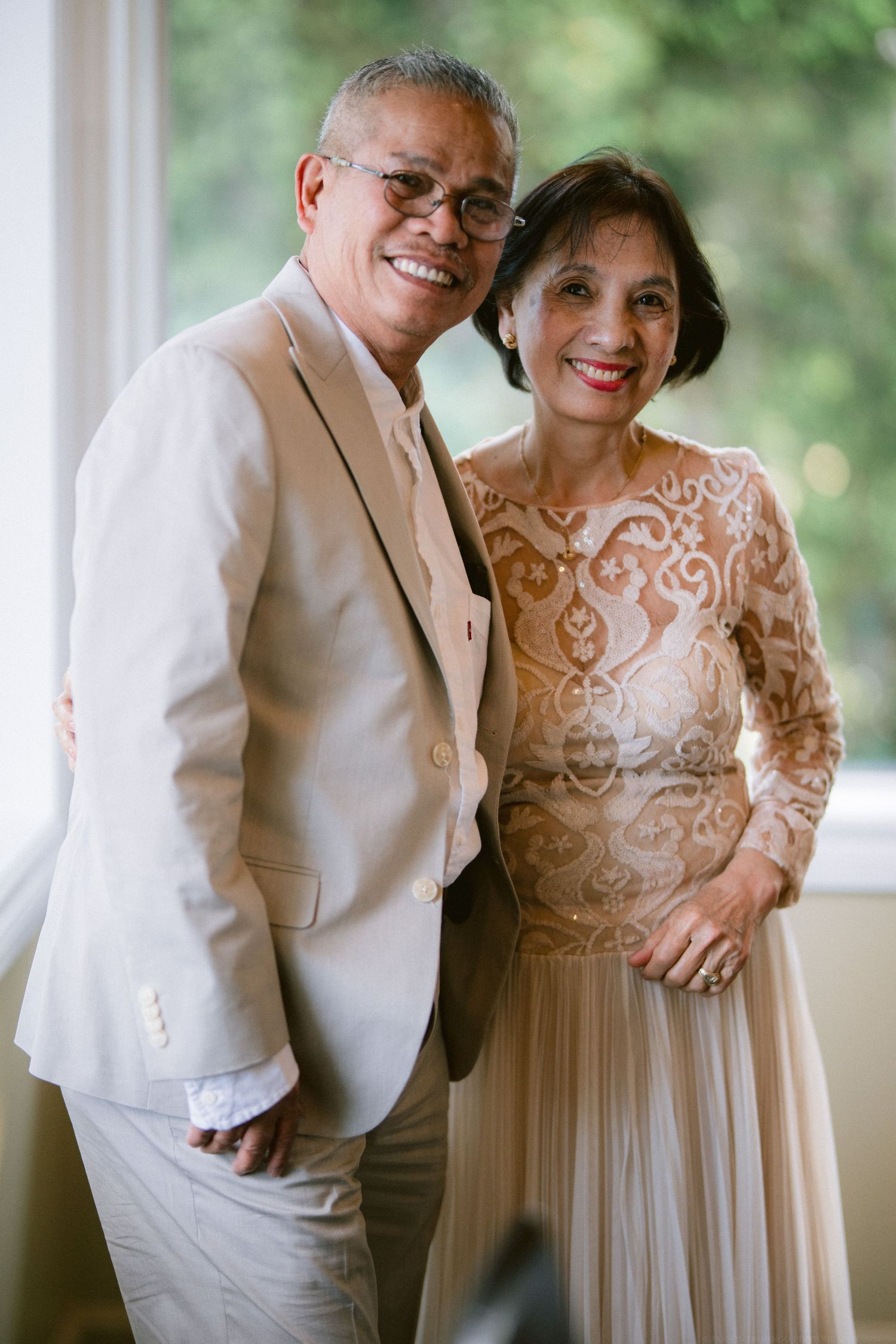 An elegant mature couple poses together, wearing formal attire with a beige suit and embroidered dress.