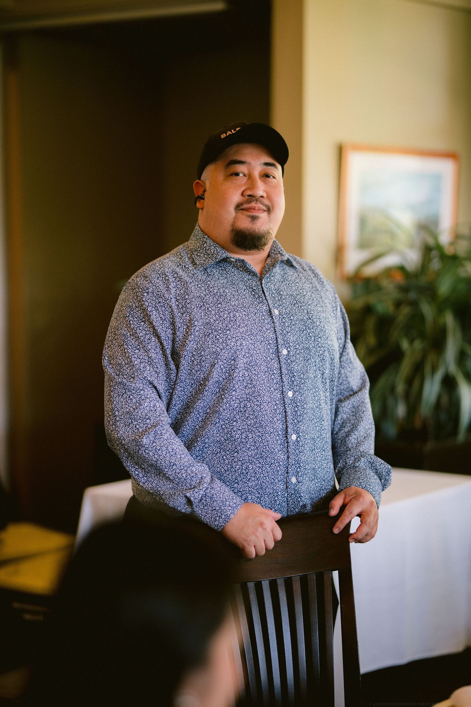 A person in a navy baseball cap and grey patterned button-down shirt stands beside a wooden chair indoors.