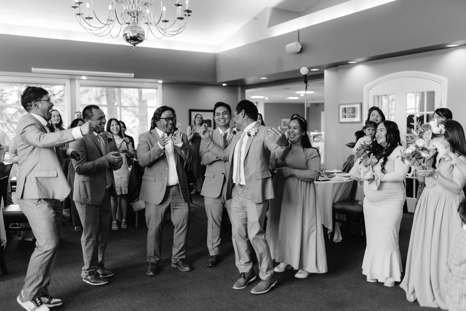 Wedding guests dance and celebrate on the dance floor at a reception venue with a chandelier overhead.