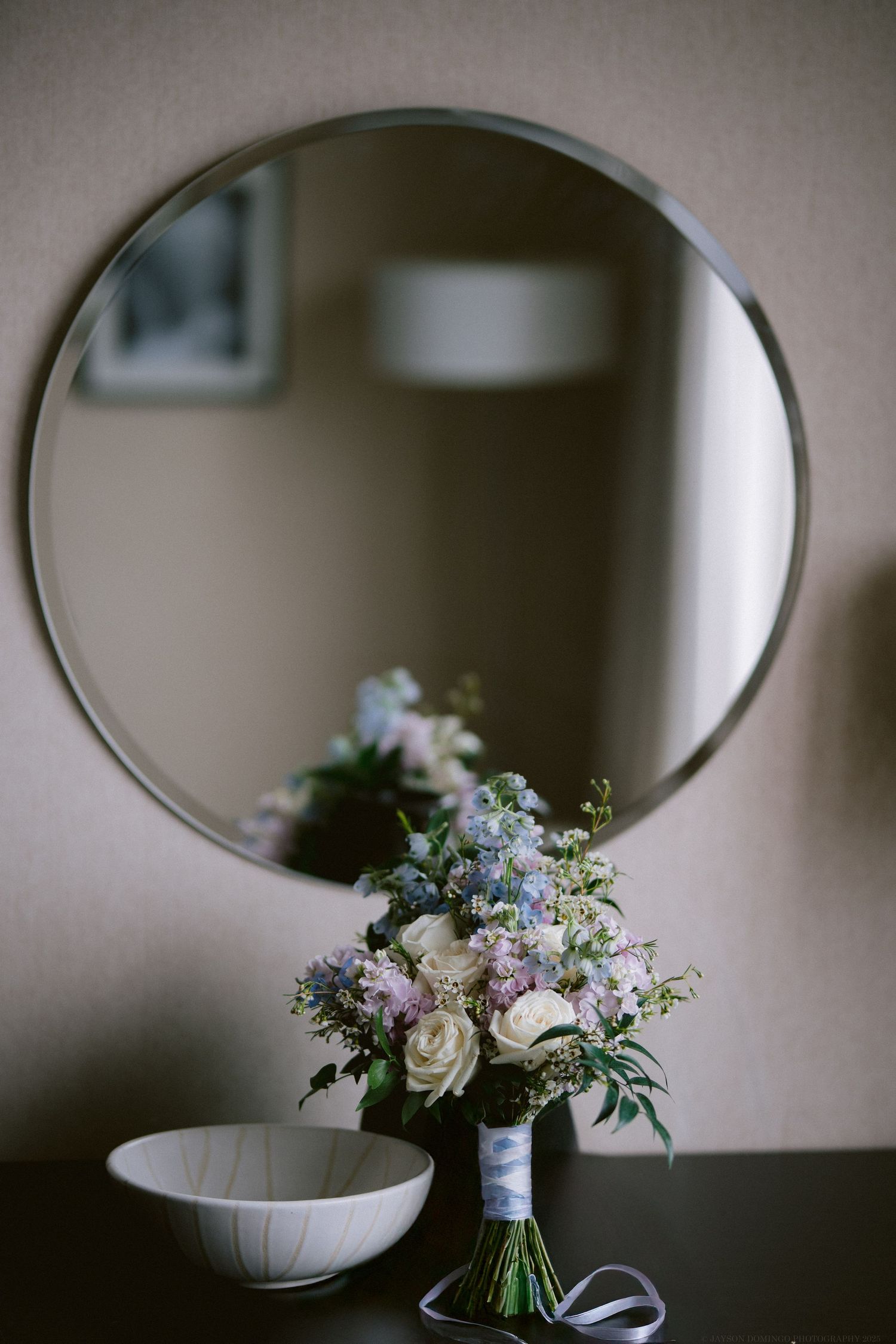 An elegant wedding bouquet with white roses and purple flowers sits beneath a round mirror.