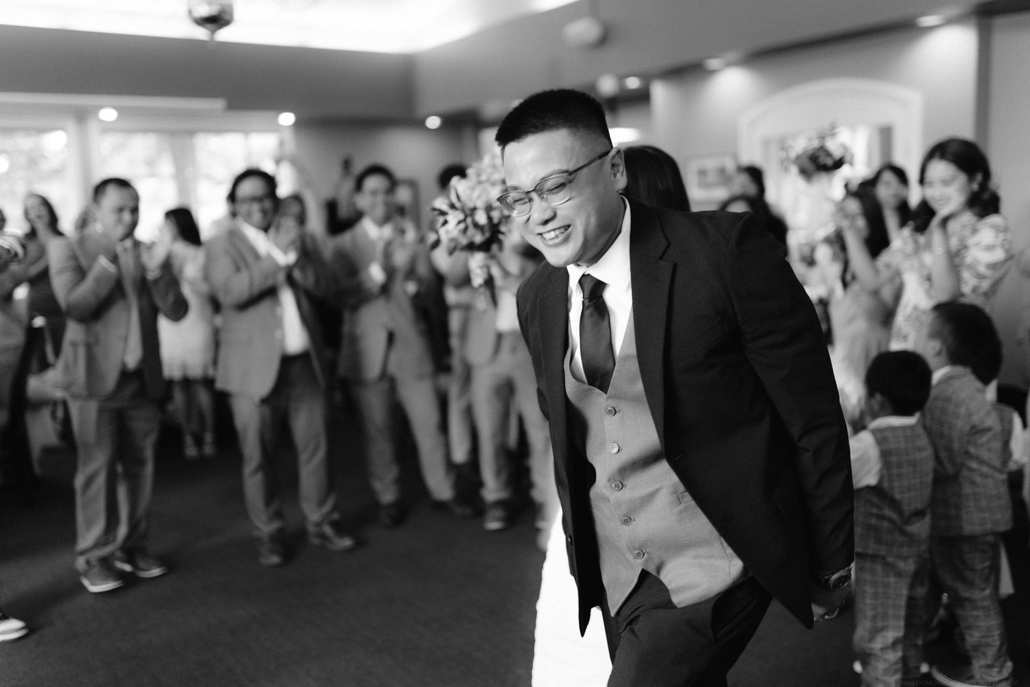 The wedding guest dances joyfully wearing a suit and vest while others watch and applaud in the background.