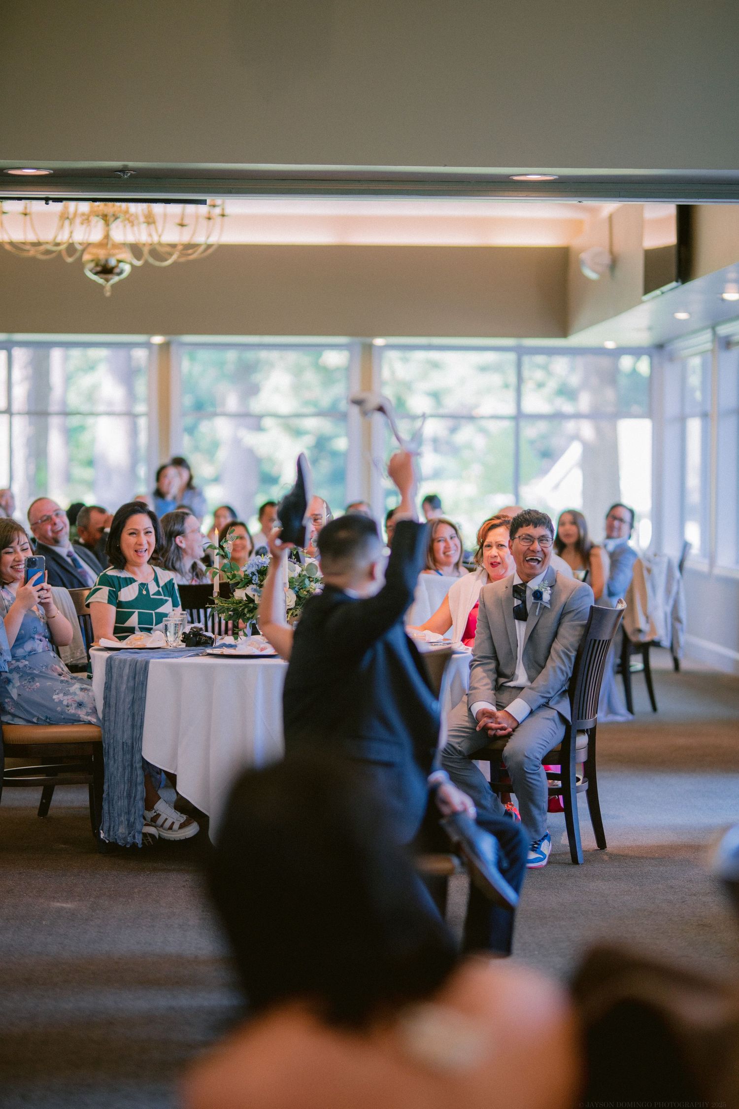 Wedding guests seated at round tables in a bright reception venue react to a speech.