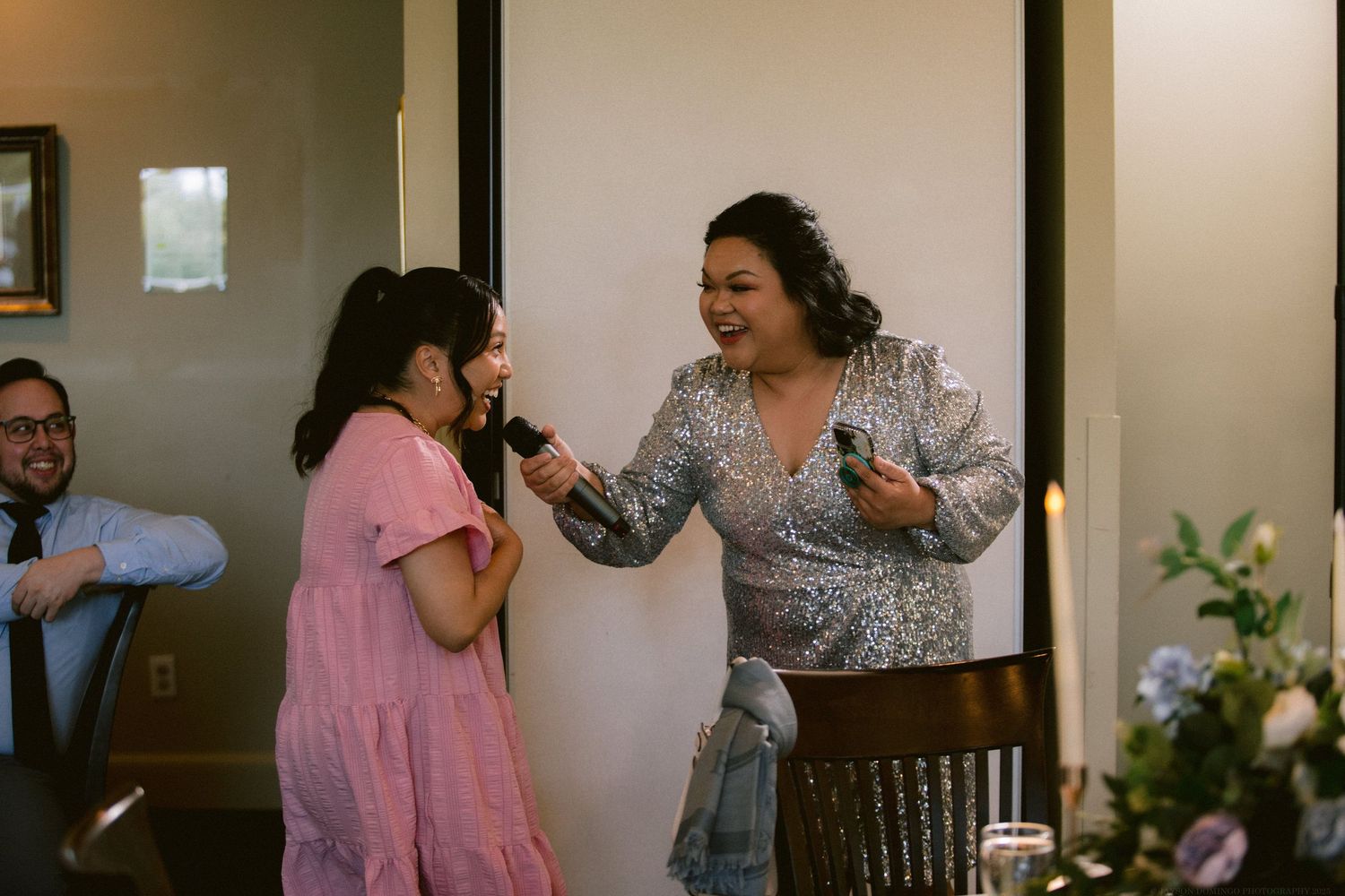 Two people share an emotional moment during an indoor gathering while someone looks on from behind.