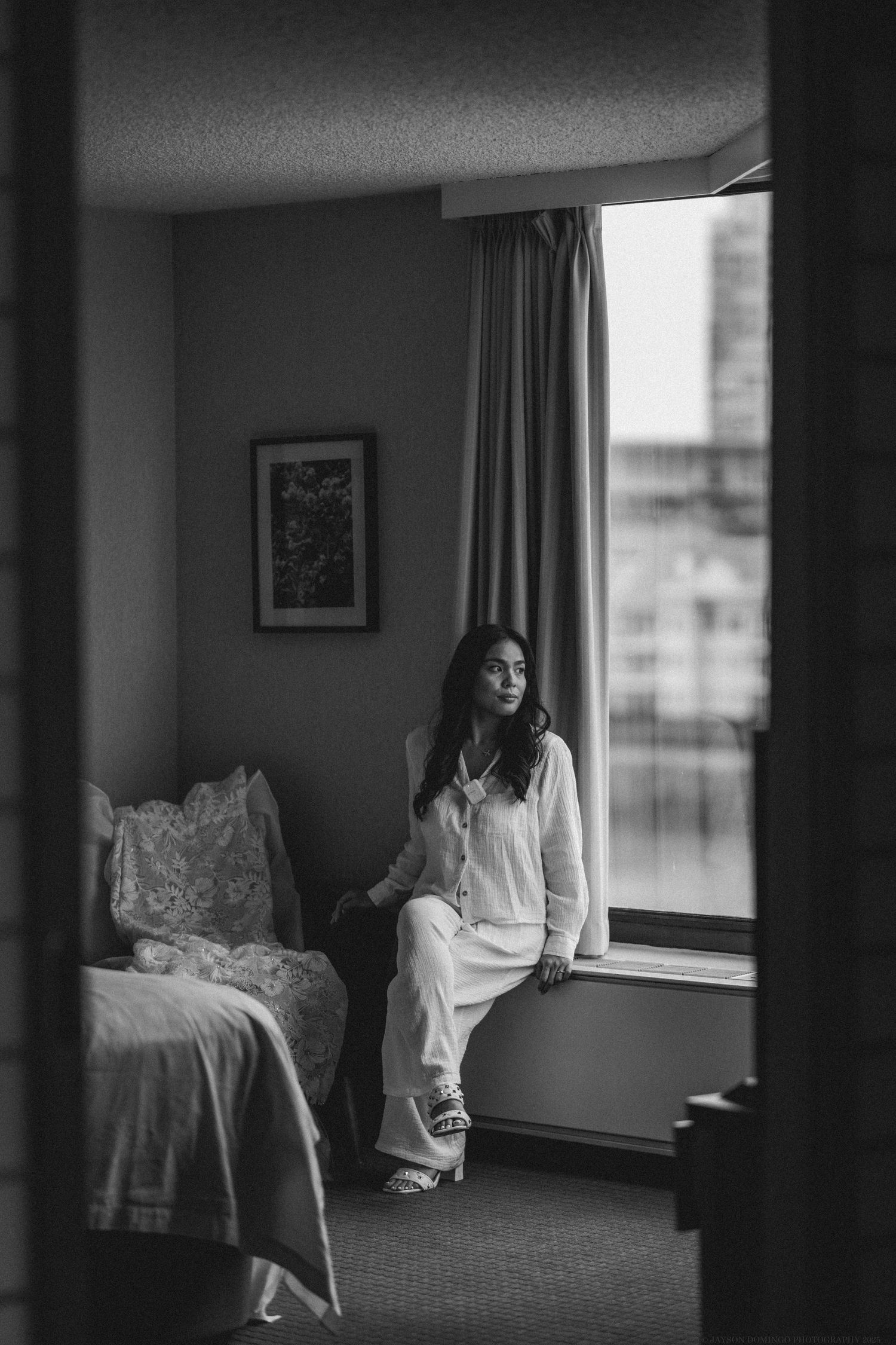Black and white photo of someone sitting pensively by a window in a hotel room.