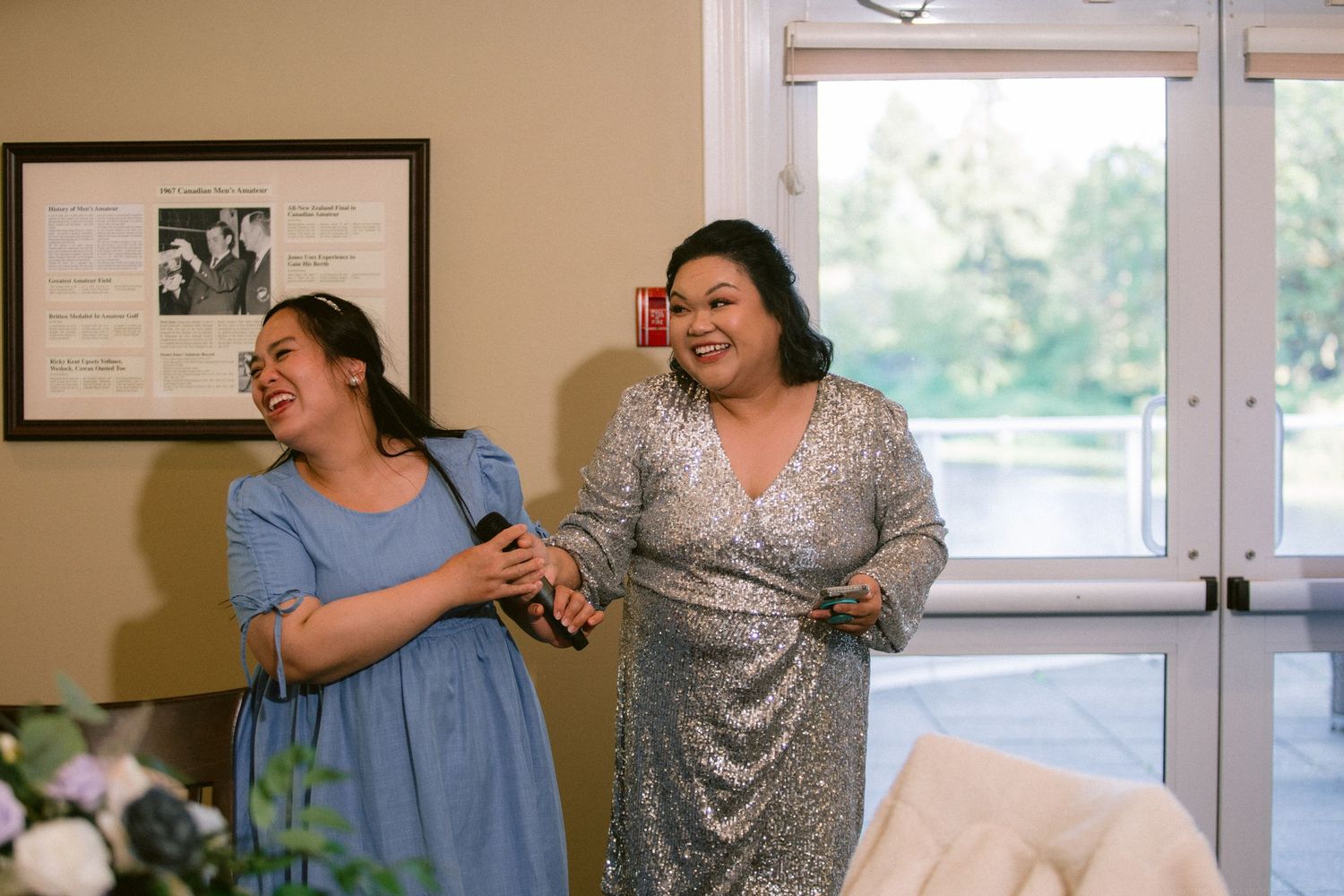 Two people in dresses share a candid laughing moment together in a bright interior room.