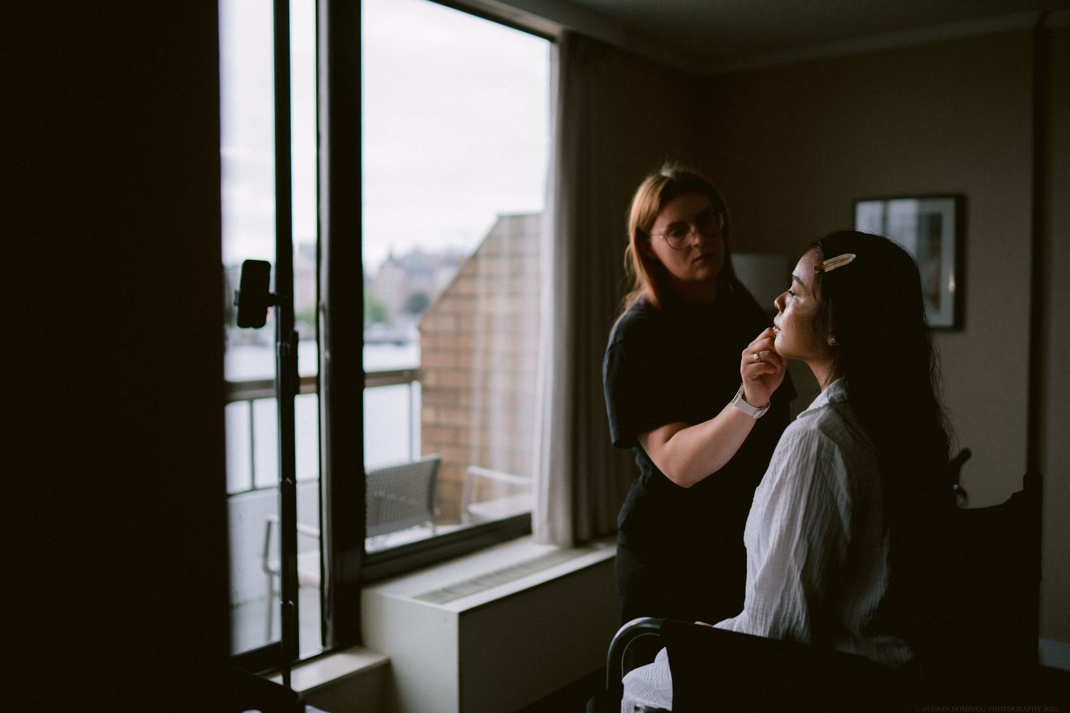 A hairstylist works on styling the bride's hair by a window.