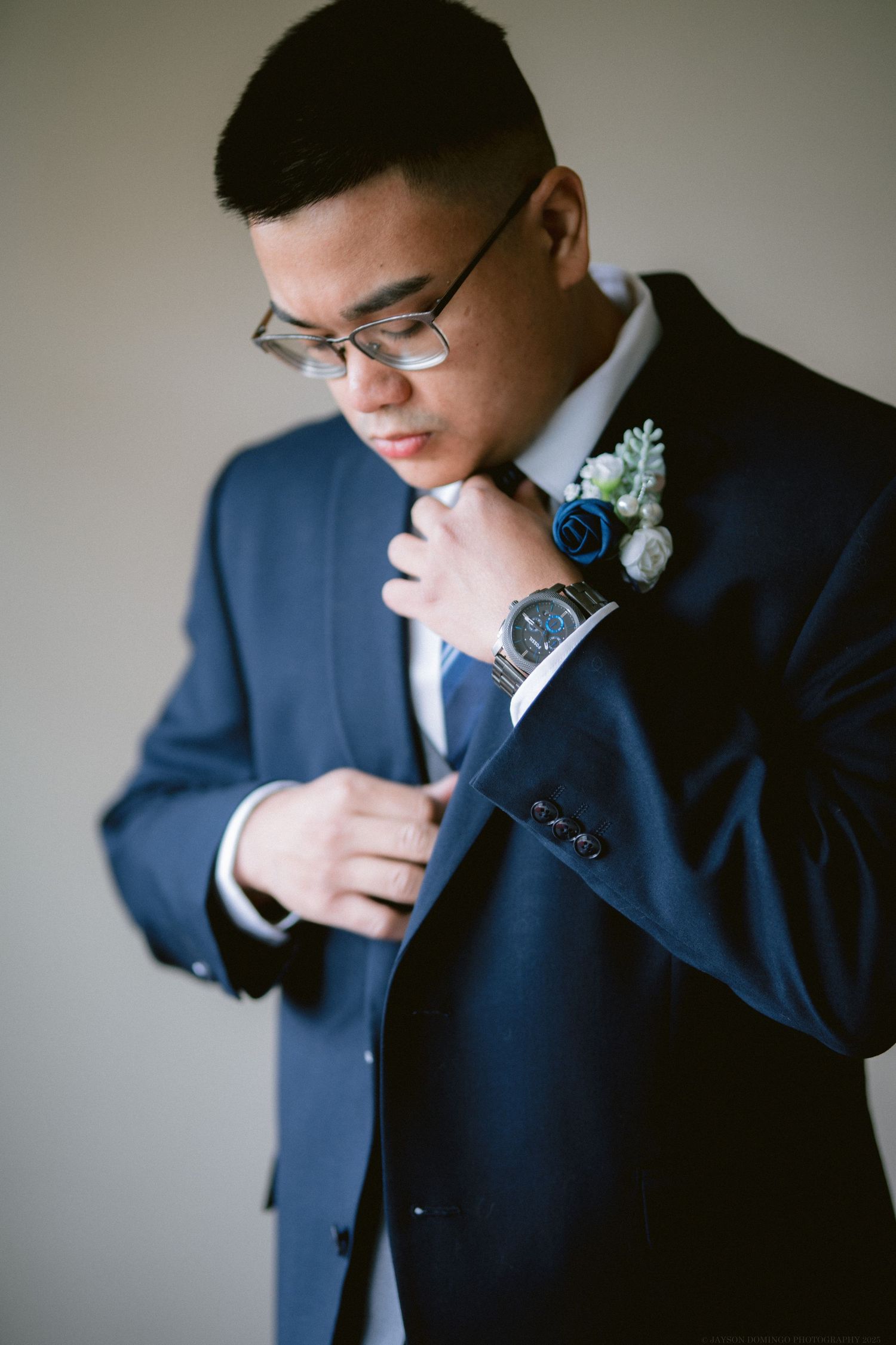 A groom in a navy suit adjusts his tie while wearing a blue boutonniere.