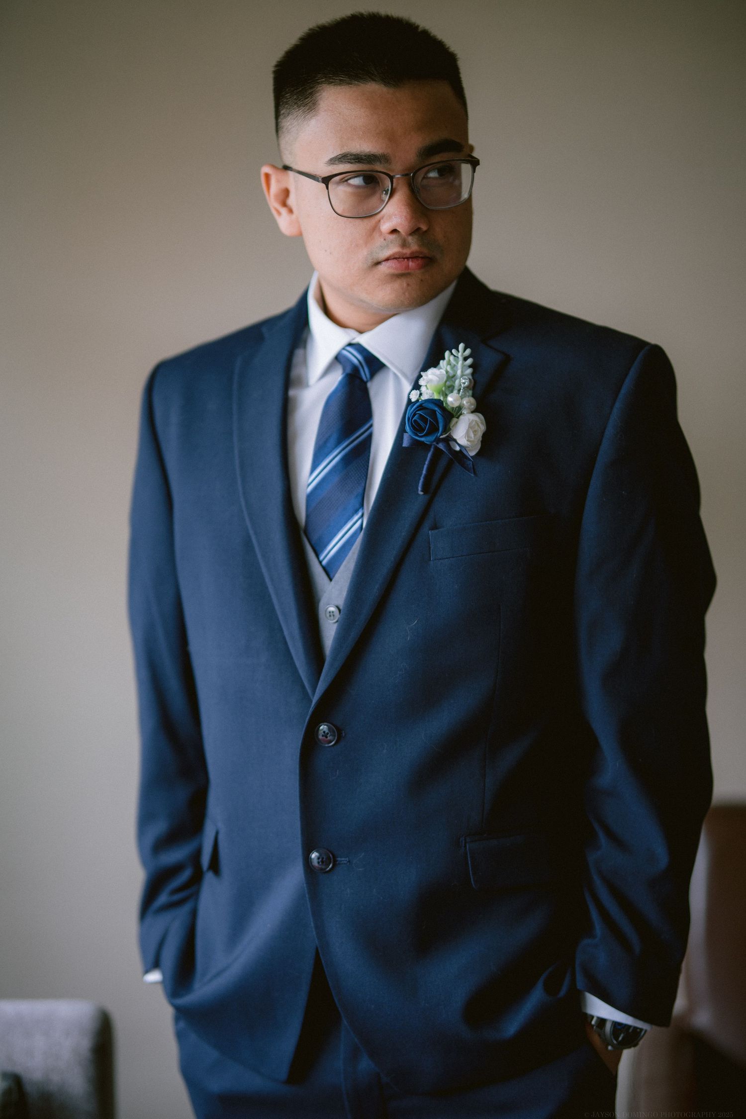 A groom in a navy blue suit with striped tie and boutonniere poses against a neutral background.