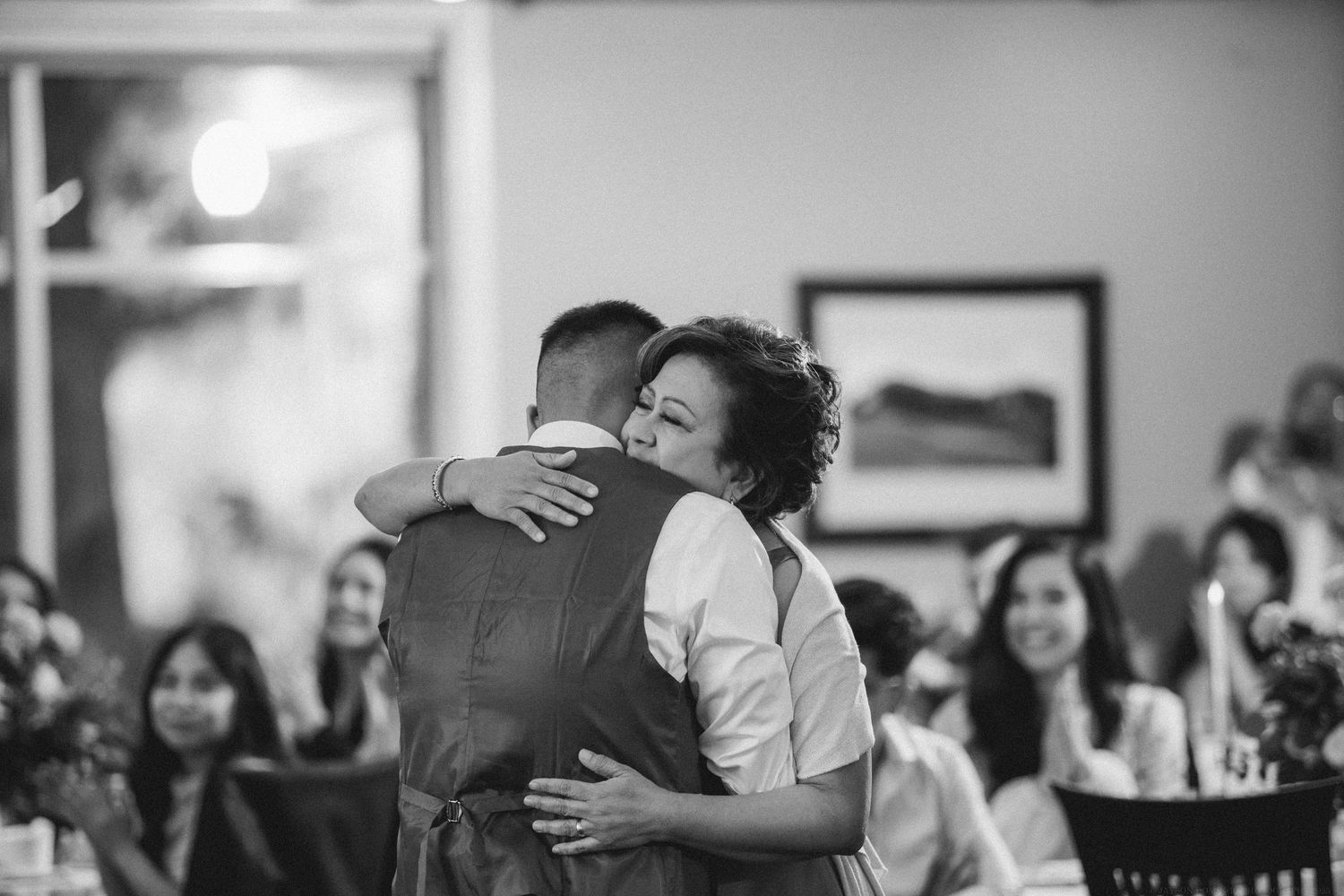 Emotional embrace captured in black and white during wedding reception with guests looking on.