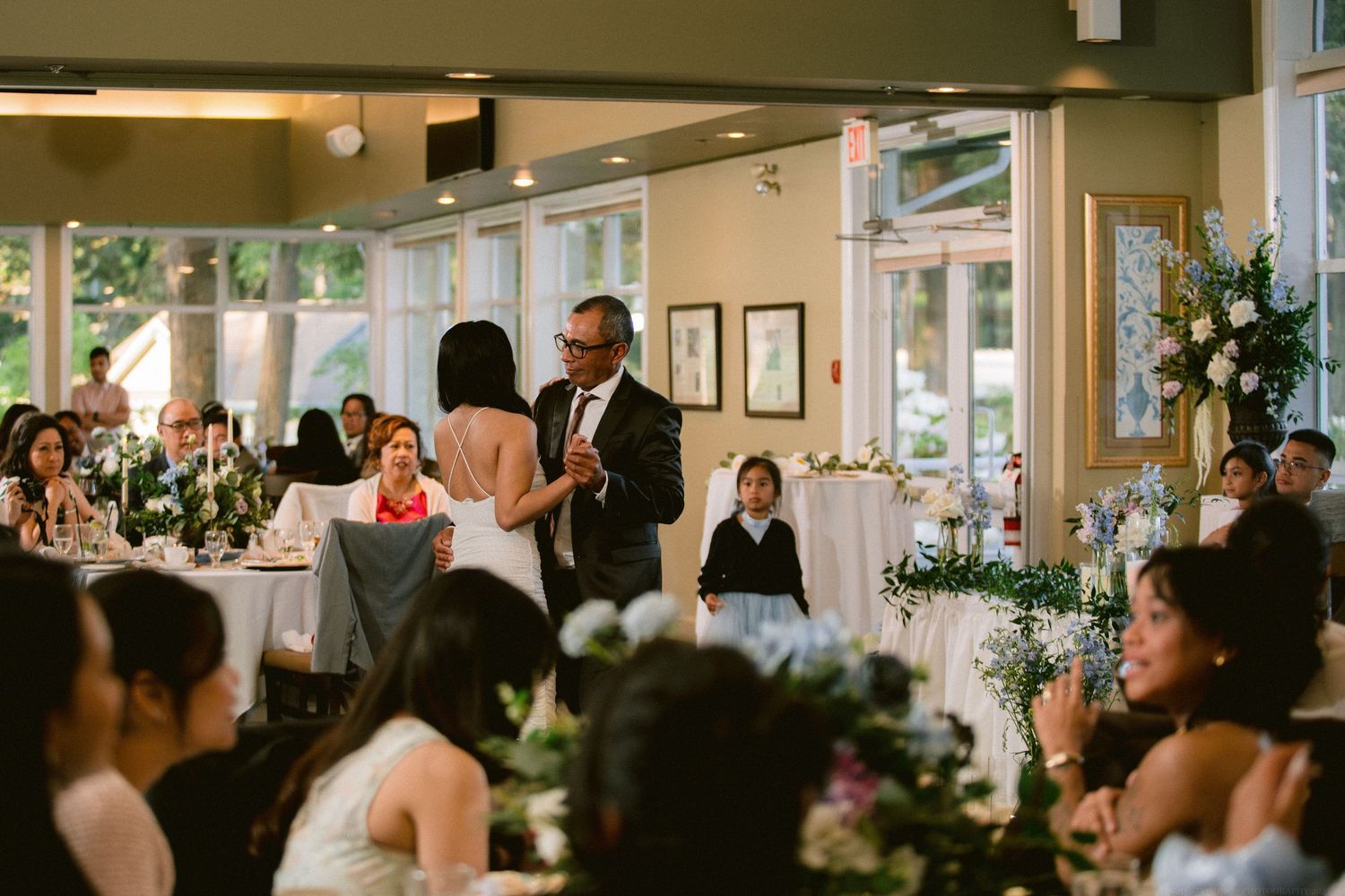 Guests watch attentively during a reception speech in an elegant venue with large windows.