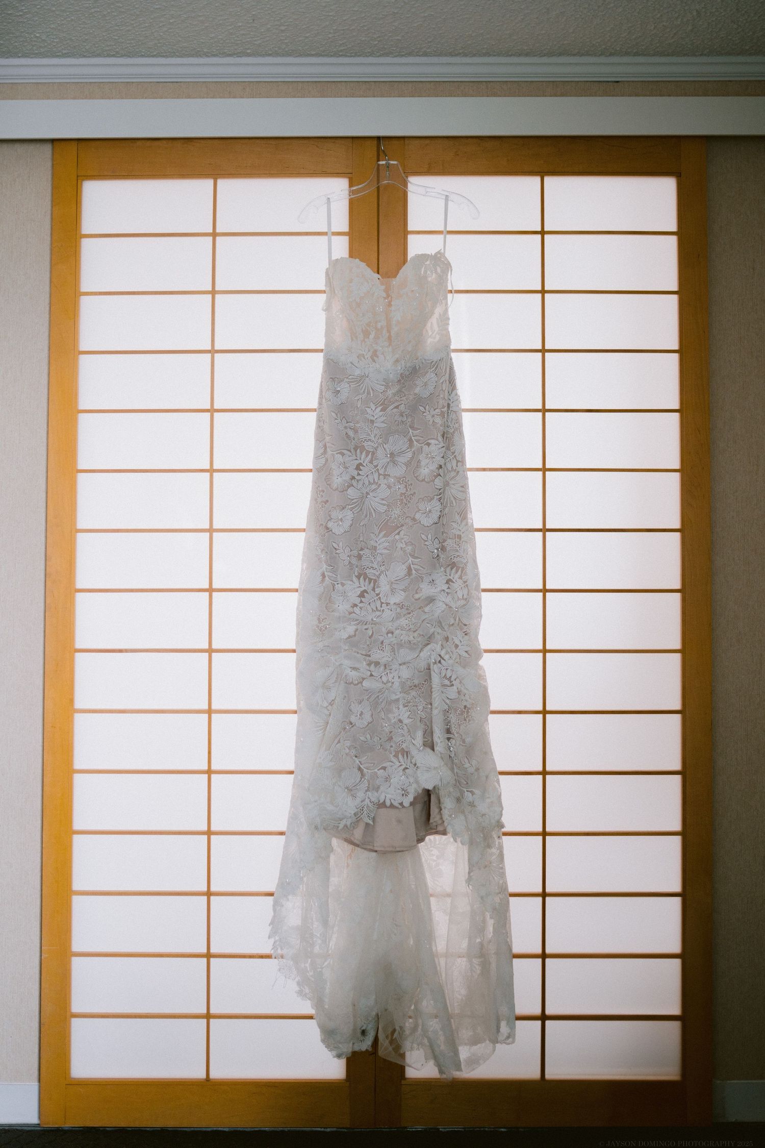 A delicate white lace wedding dress hangs on a wooden door with traditional Japanese shoji screen panels.