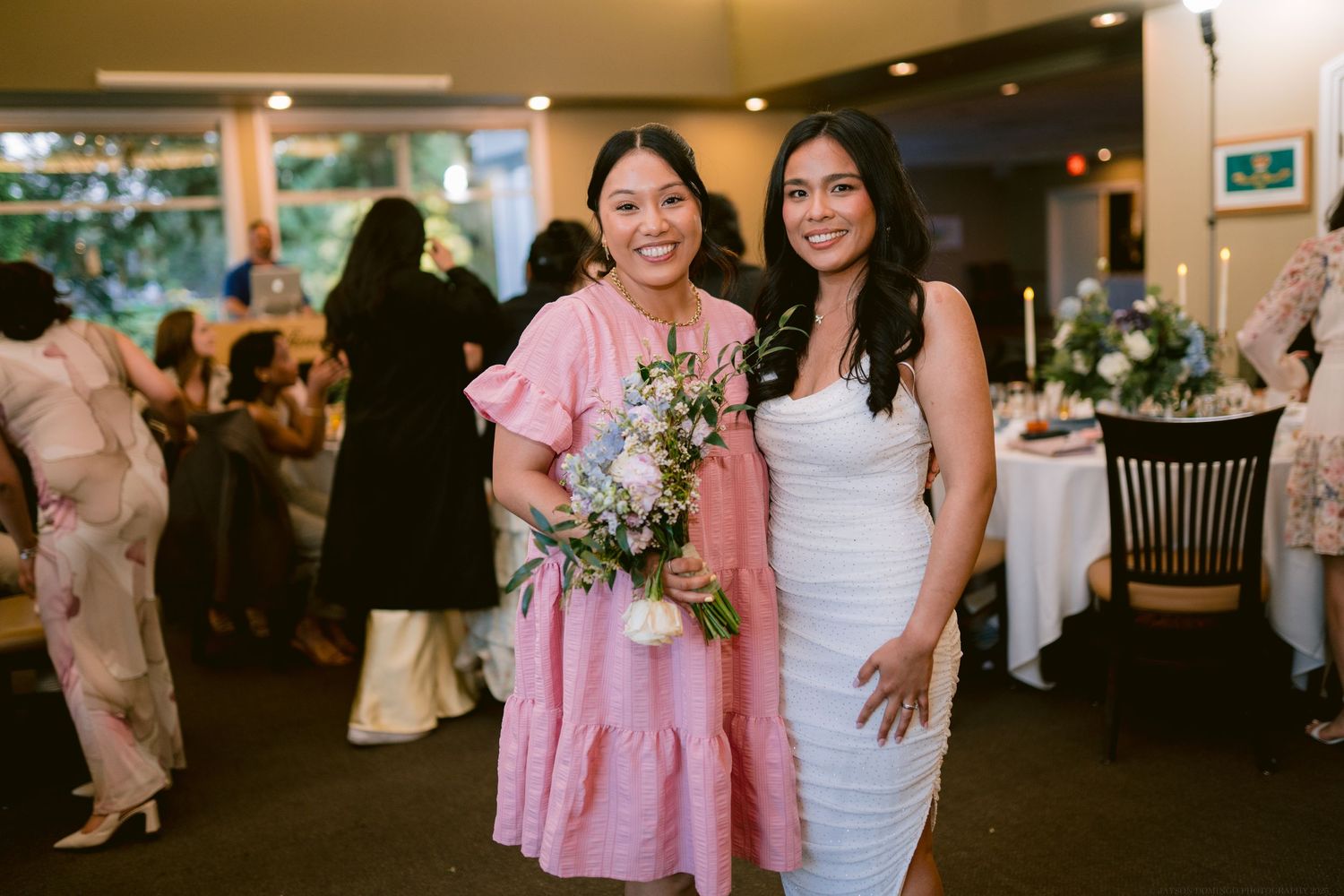Two women at a wedding reception, one in pink dress holding flowers and one in white dress.