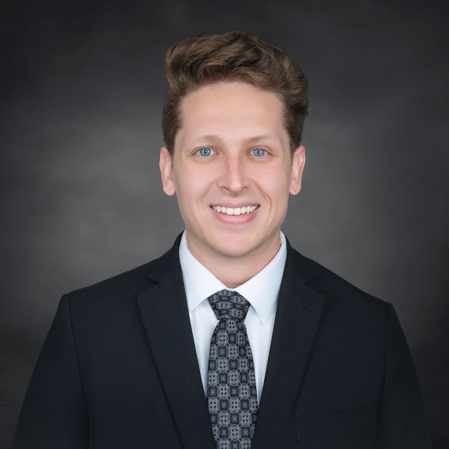 Professional headshot of someone in a dark suit with patterned tie against a gray backdrop.