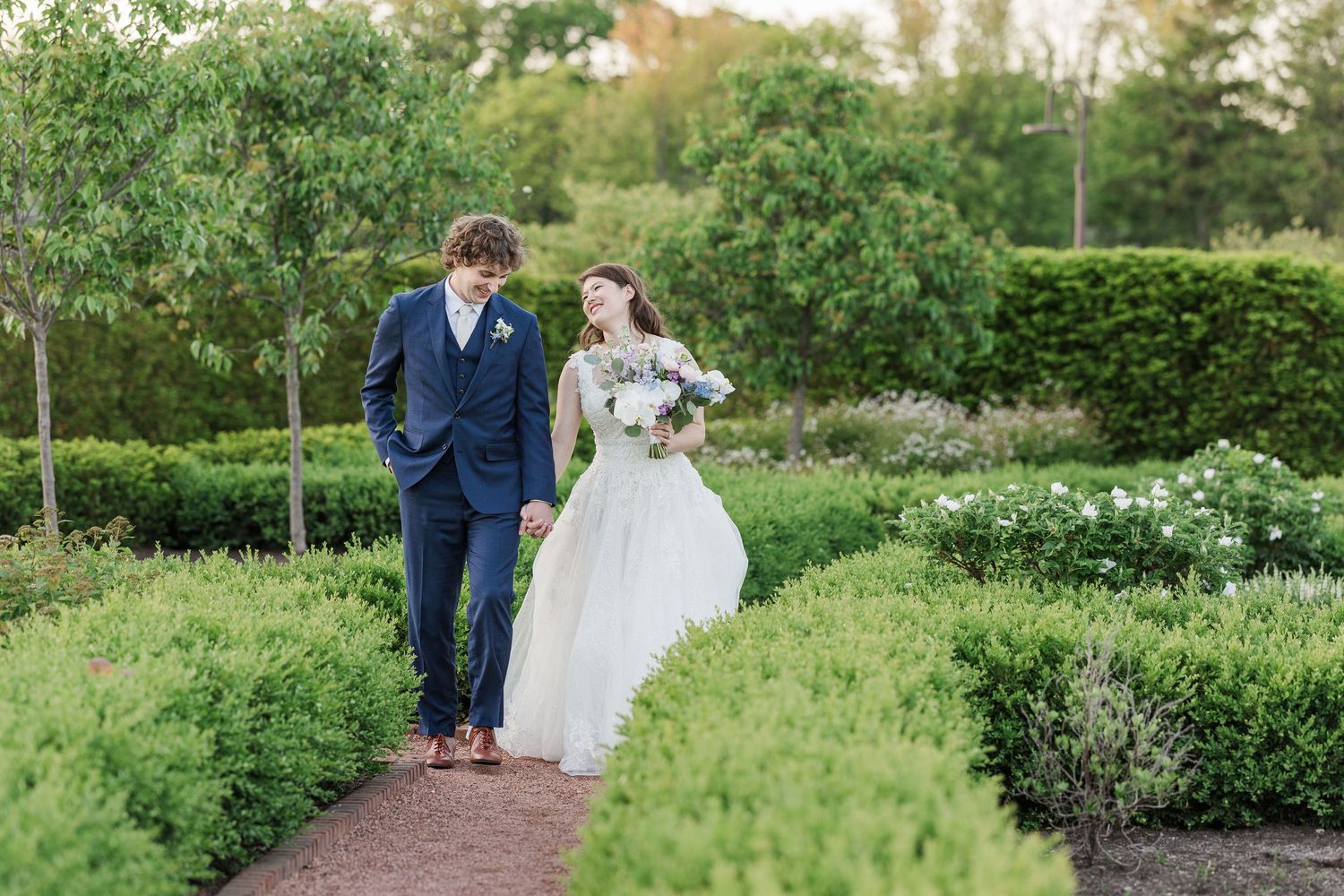 A romantic garden path emerges through lush greenery with a couple taking a stroll in formal attire.