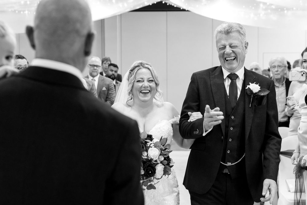 Bride walking down the aisle with her father, both laughing with joy at the Village Hotel, Portsmouth.