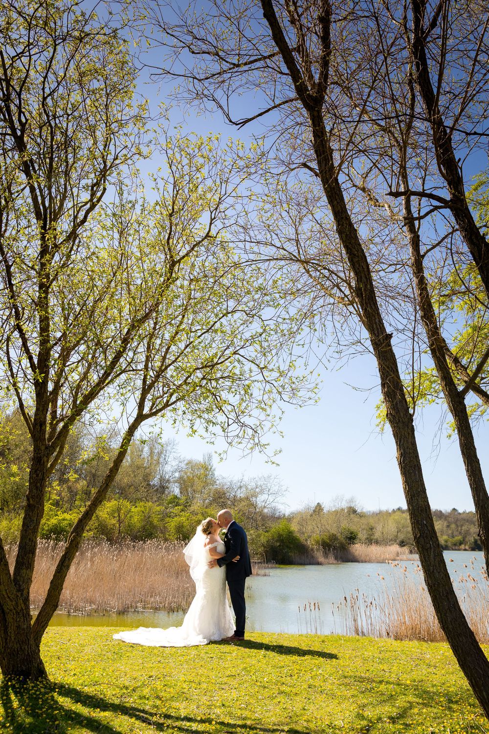 Bride and groom kissing under trees by the lake near the Village Hotel, Portsmouth, in beautiful spring sunlight.