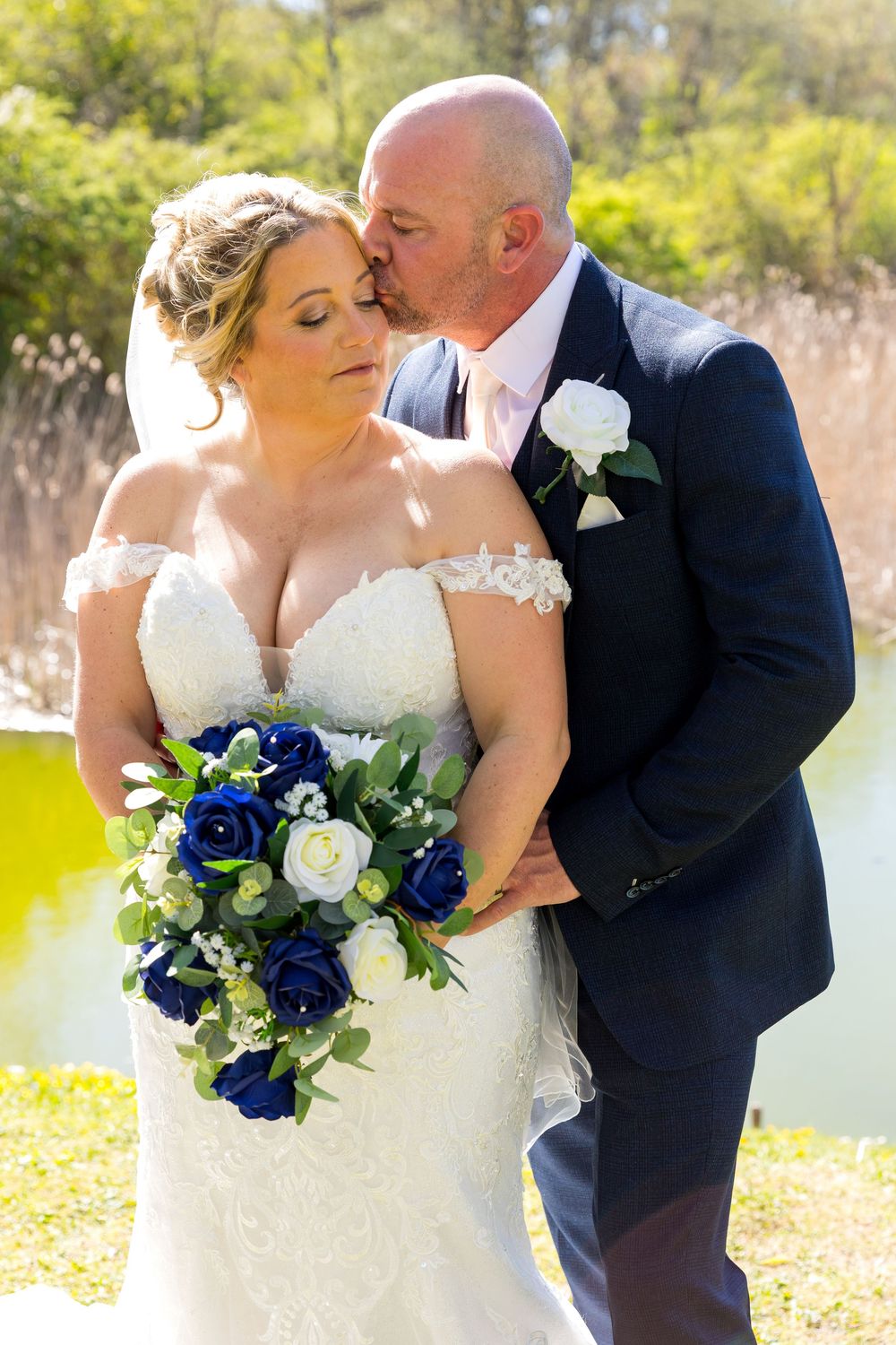 Bride and groom embracing by the water, groom kisses bride's forehead on a sunny wedding day.