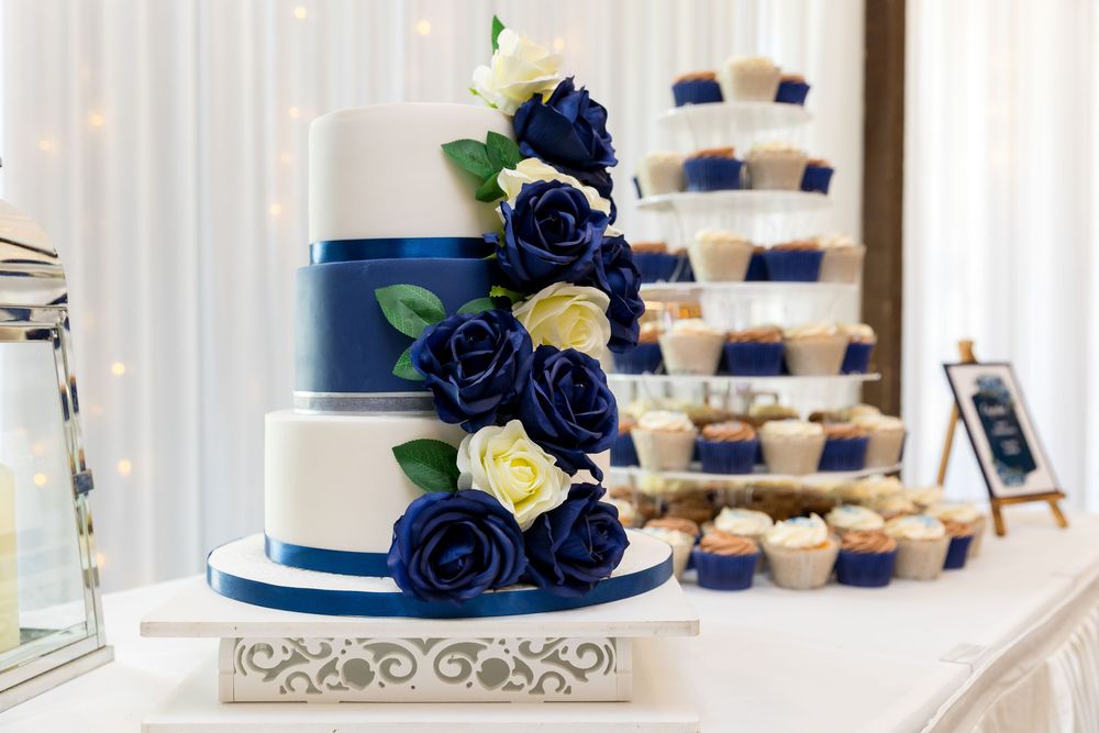 Tiered white wedding cake with navy and ivory rose decorations, next to a tower of cupcakes.