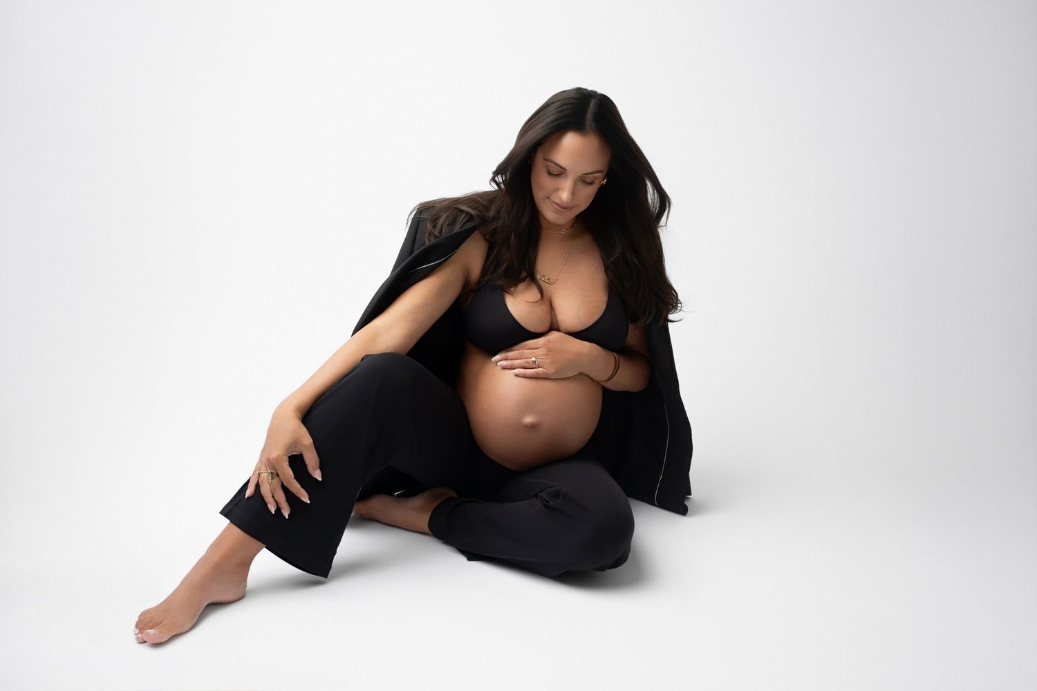 Expectant mother poses in black outfit against minimalist white backdrop.