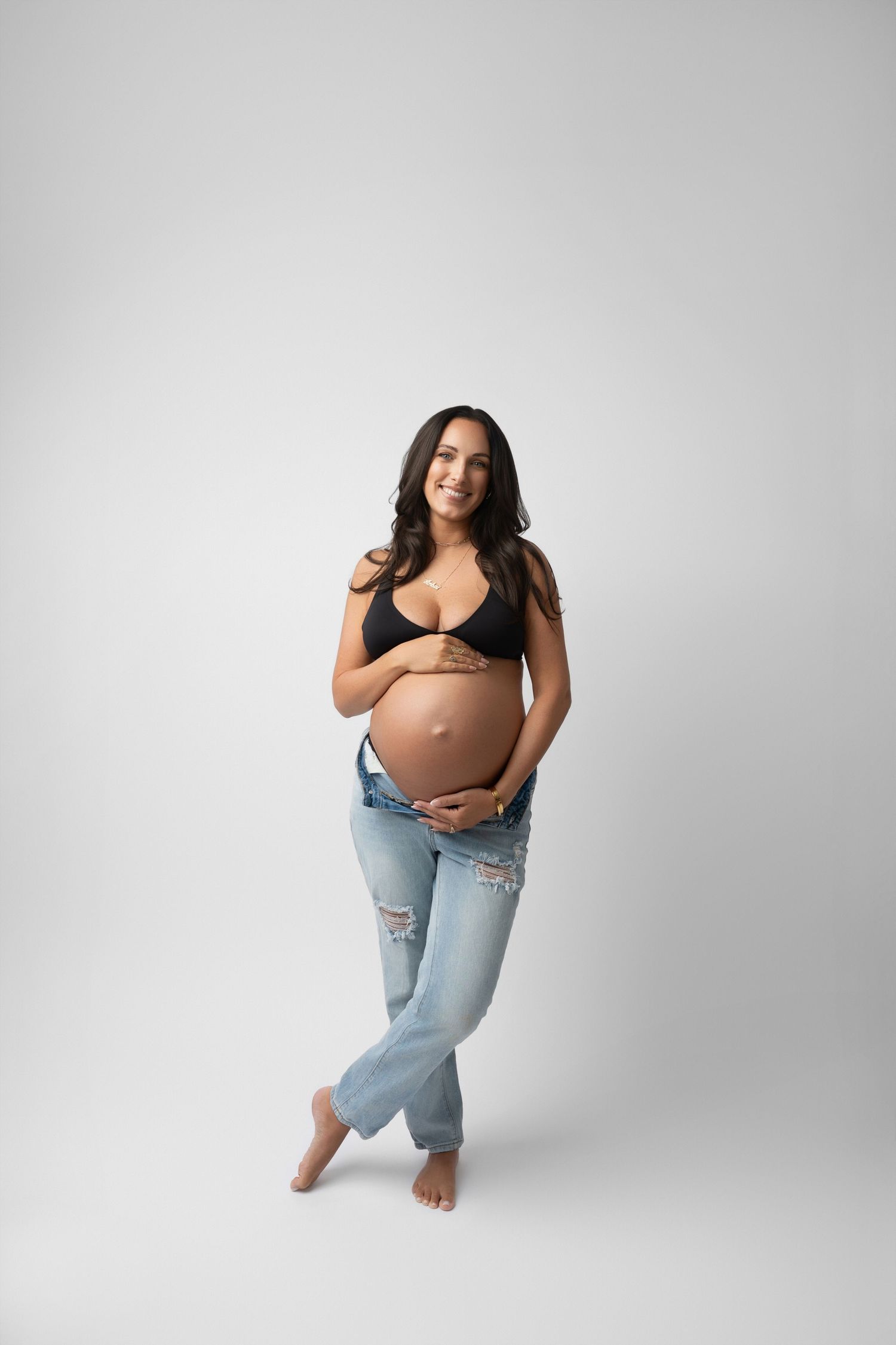 Casual maternity portrait with ripped jeans and black top against white background.