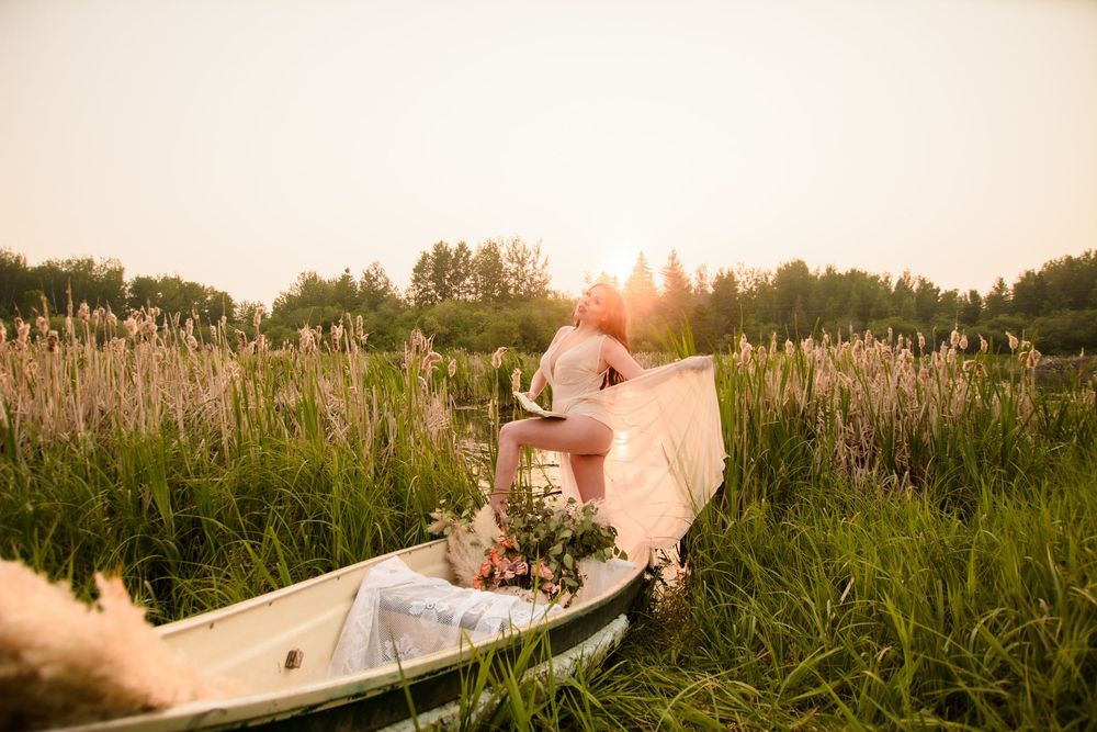 Person in flowing dress sits reading in vintage rowboat surrounded by tall cattails at sunset in dreamy summer meadow.