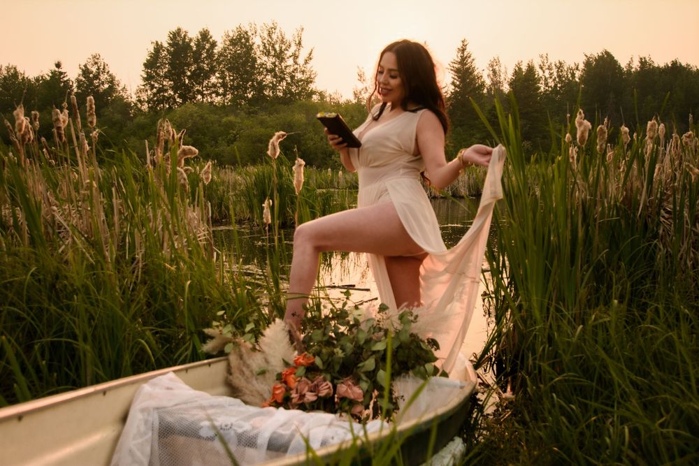 A dreamy sunset scene with a person in a flowing dress standing in a boat among tall reeds and wildflowers.
