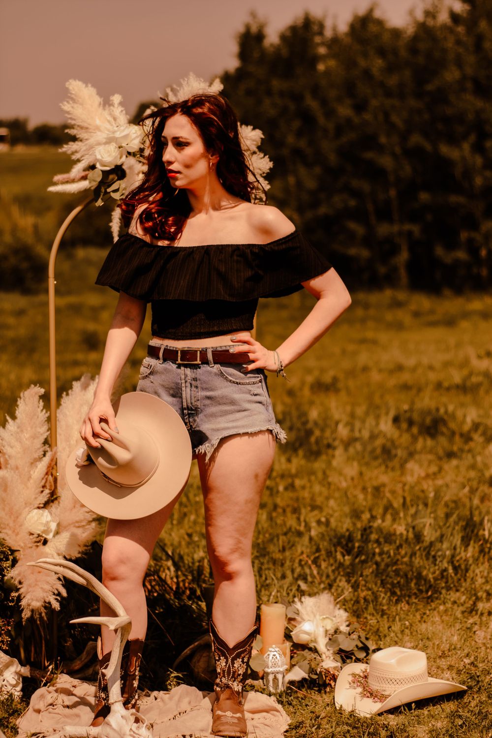 A rustic outdoor scene with someone in casual clothing sitting in a meadow surrounded by pampas grass and decorative elements.