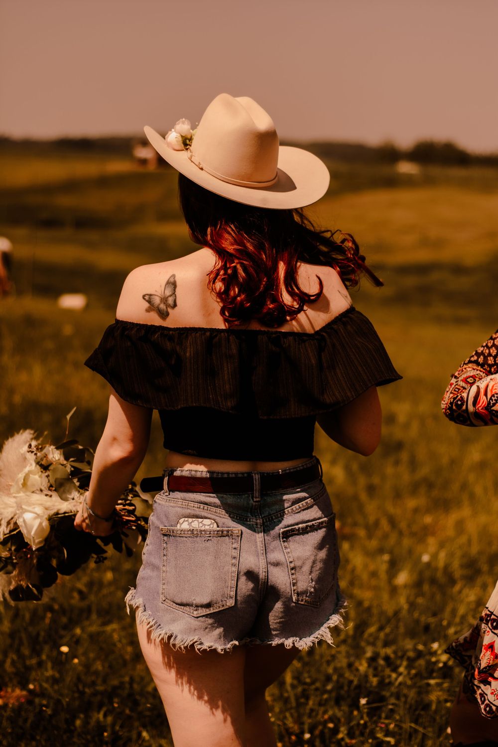 Back view of a person in denim shorts and cowboy hat against a rustic sunset backdrop.