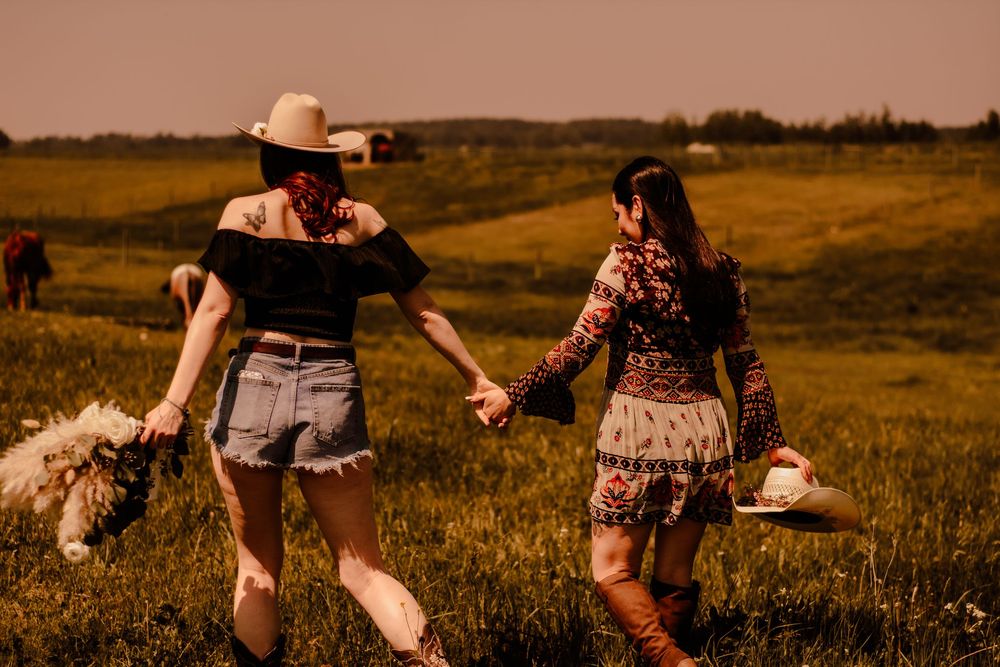 Silhouettes walk hand in hand through a golden field at sunset wearing cowboy hats and western attire.
