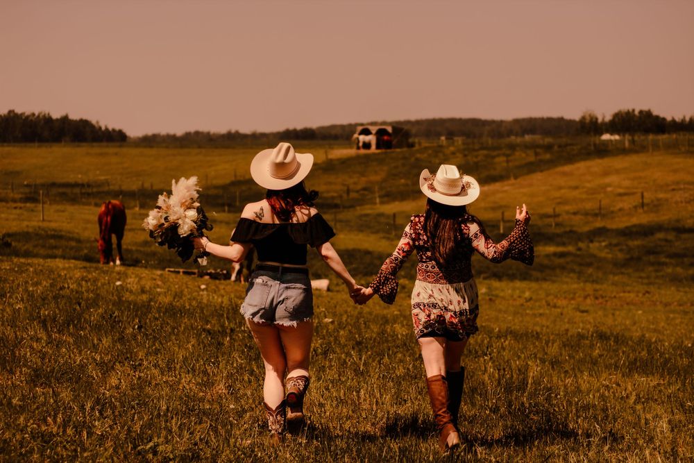 A couple in western attire strolls through a rustic field during golden hour.