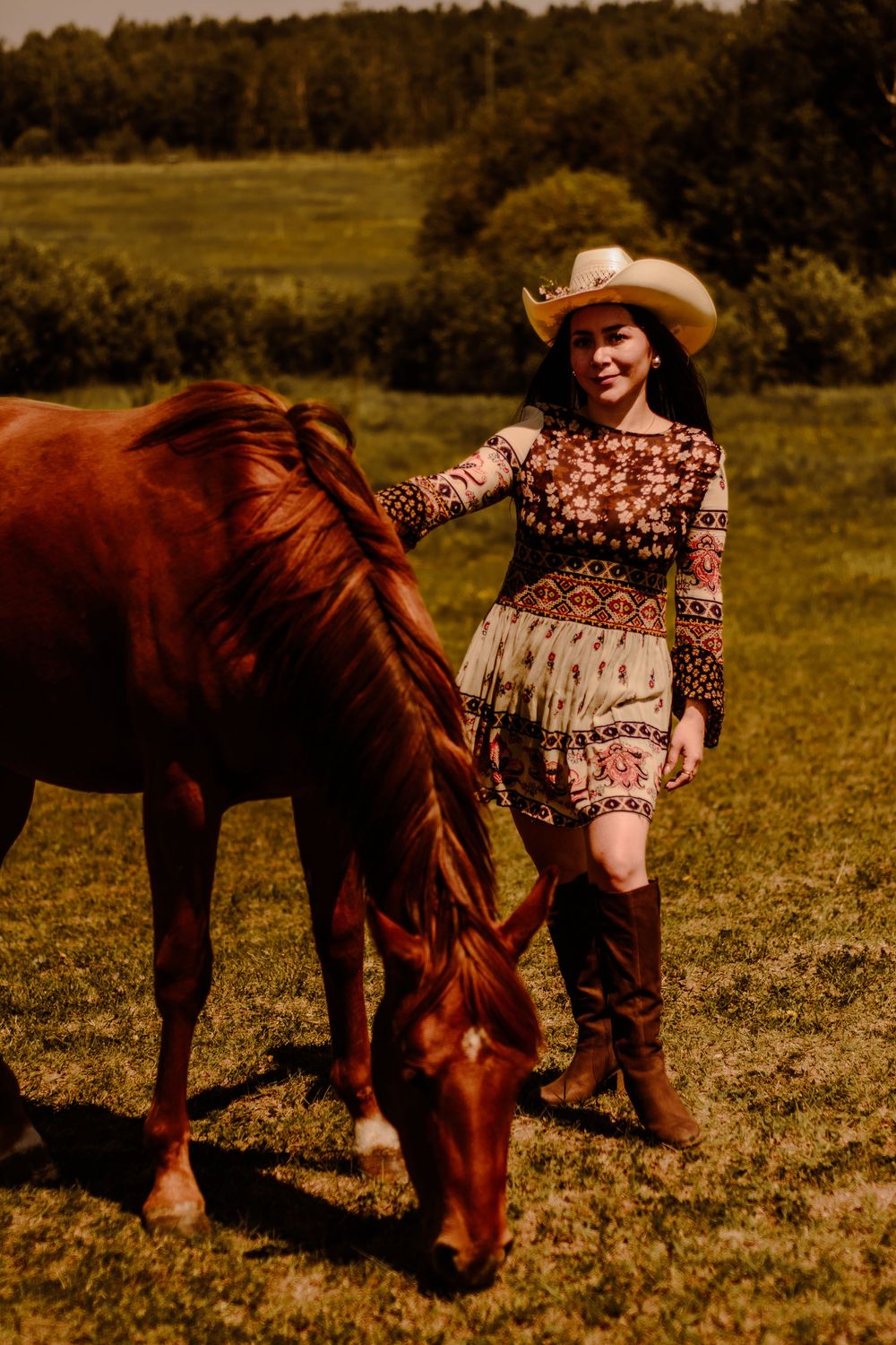 A warm-toned portrait of someone in a cowboy hat and bohemian dress petting a brown horse in a field.