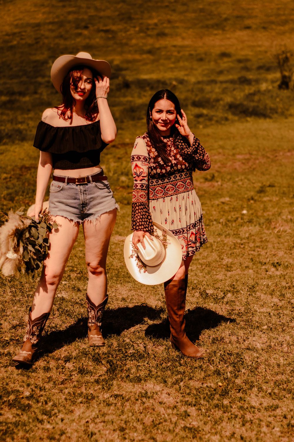Two people in cowboy boots and western attire run playfully across a grassy field at golden hour.